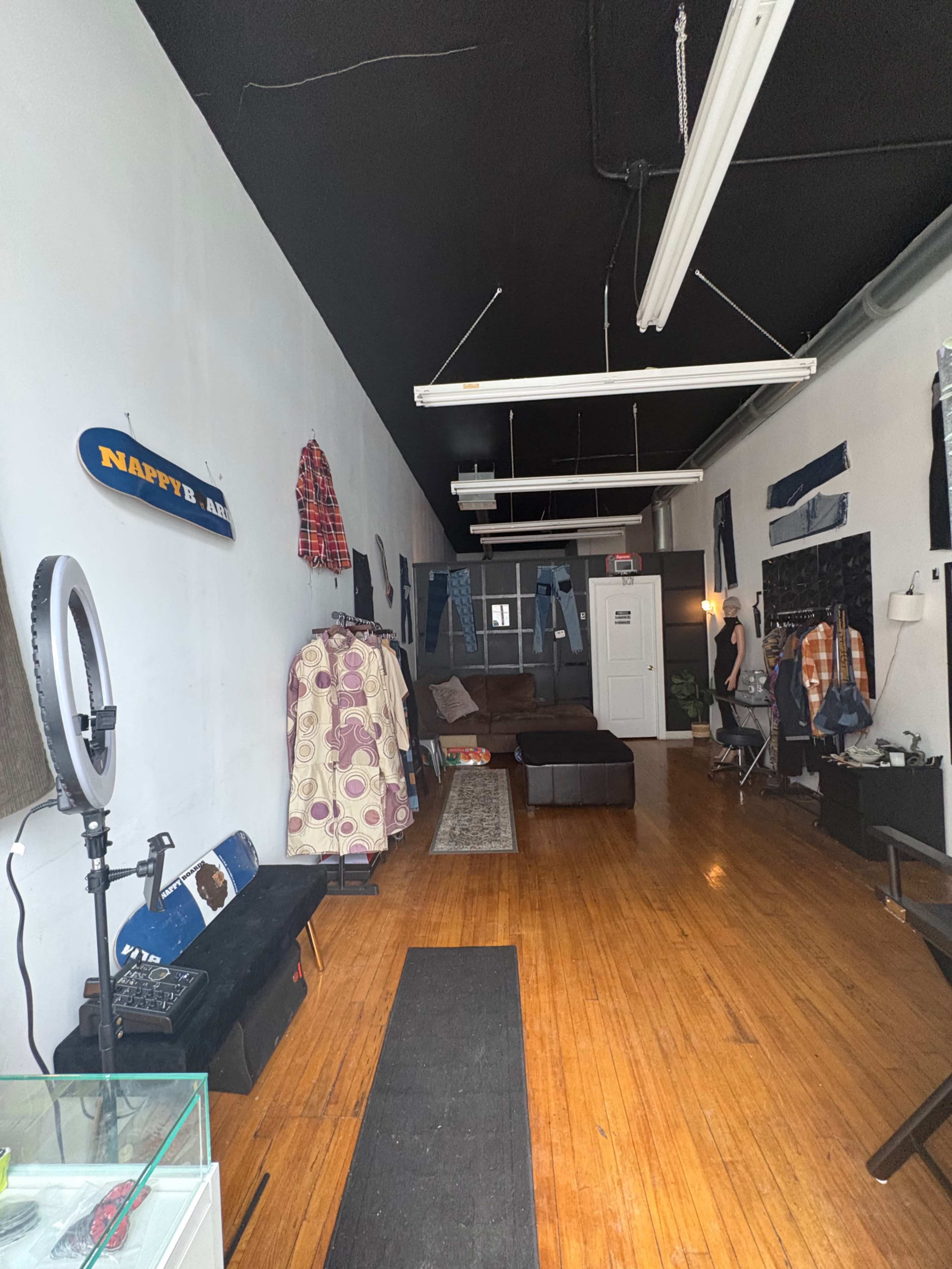 The image shows a long, narrow space with hardwood floors, featuring various clothing items displayed on racks and walls, along with a couch and a door at the back.