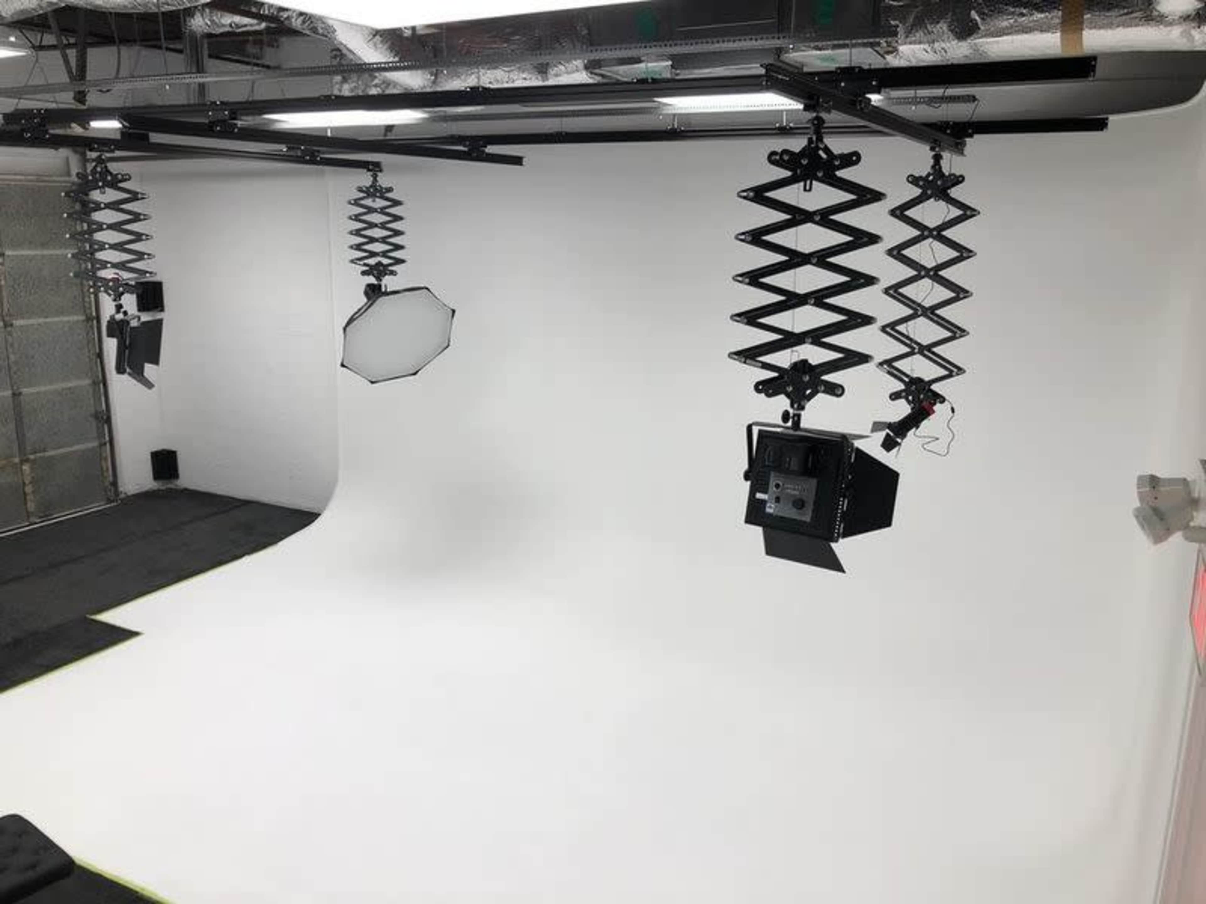 A photography studio with a white backdrop, overhead lights, and a black carpeted floor.