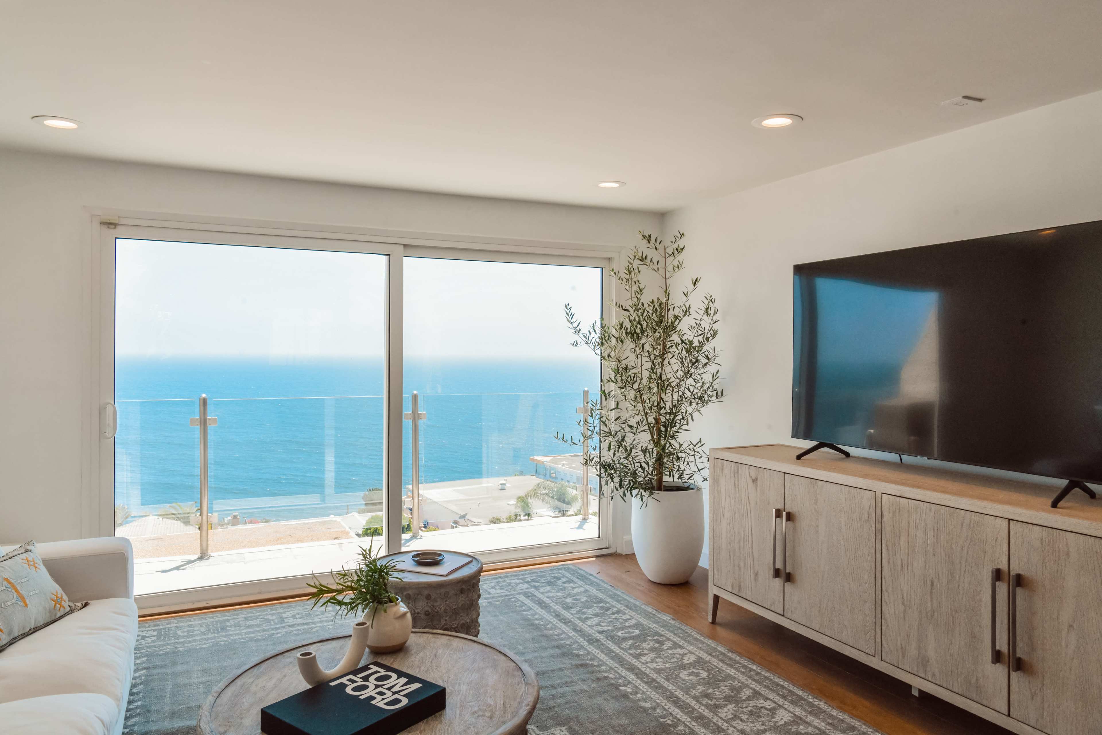 The image shows a modern living room with a large window overlooking the ocean, featuring a sofa, a TV stand, and a coffee table.