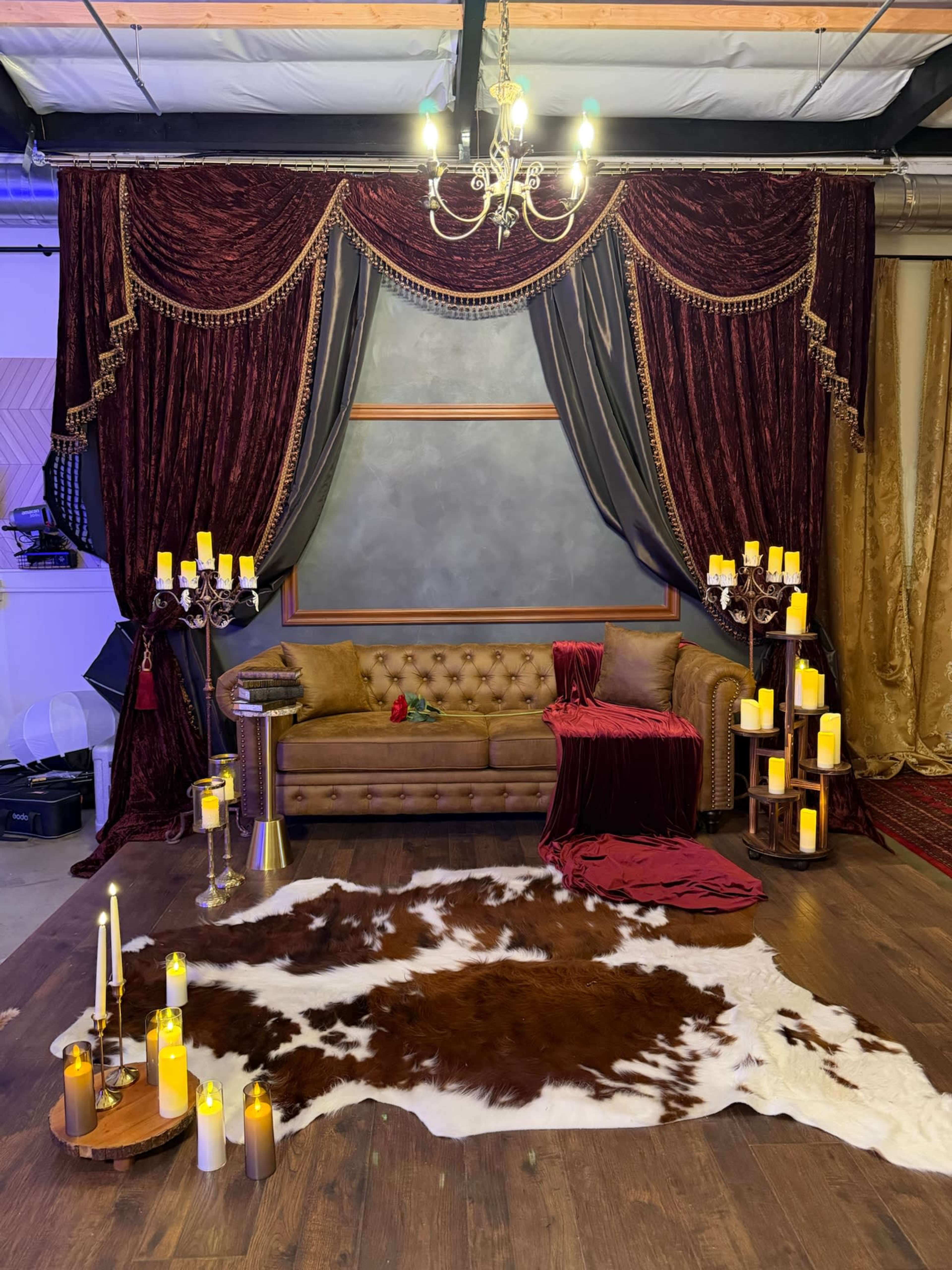 The image features a cozy interior scene with a brown tufted sofa, ornate curtains, decorative candlesticks, and a cowhide rug arranged around a dimly lit space.