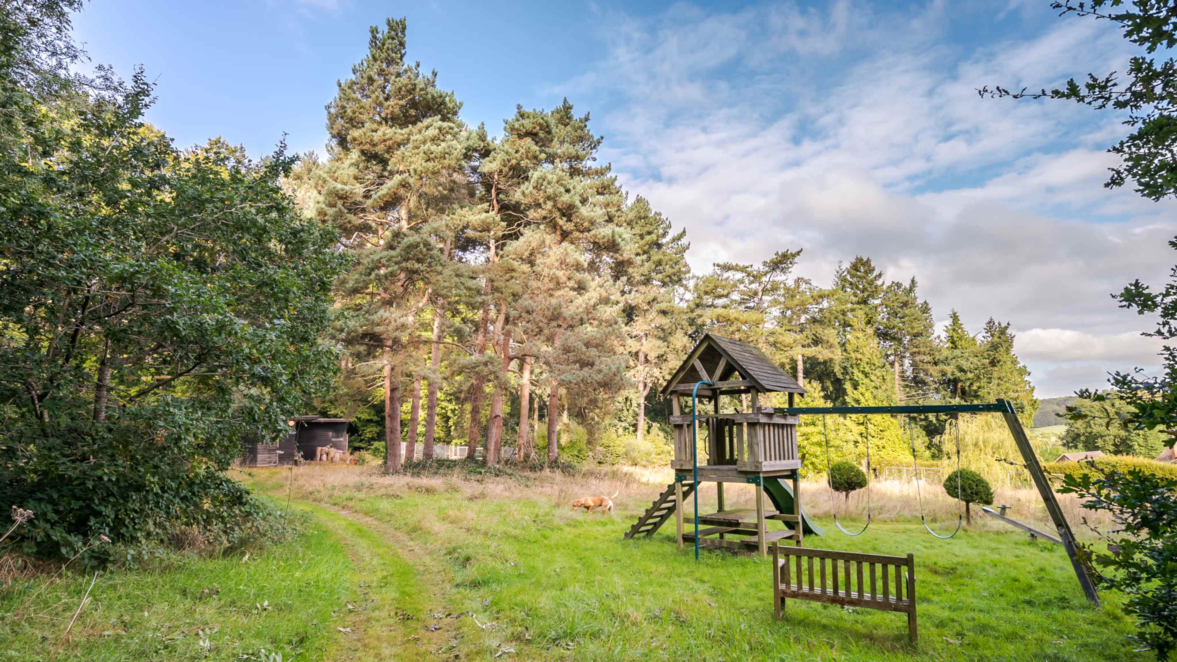 A wooden playhouse with swings is positioned near a wooded area with tall trees and a grassy pathway.
