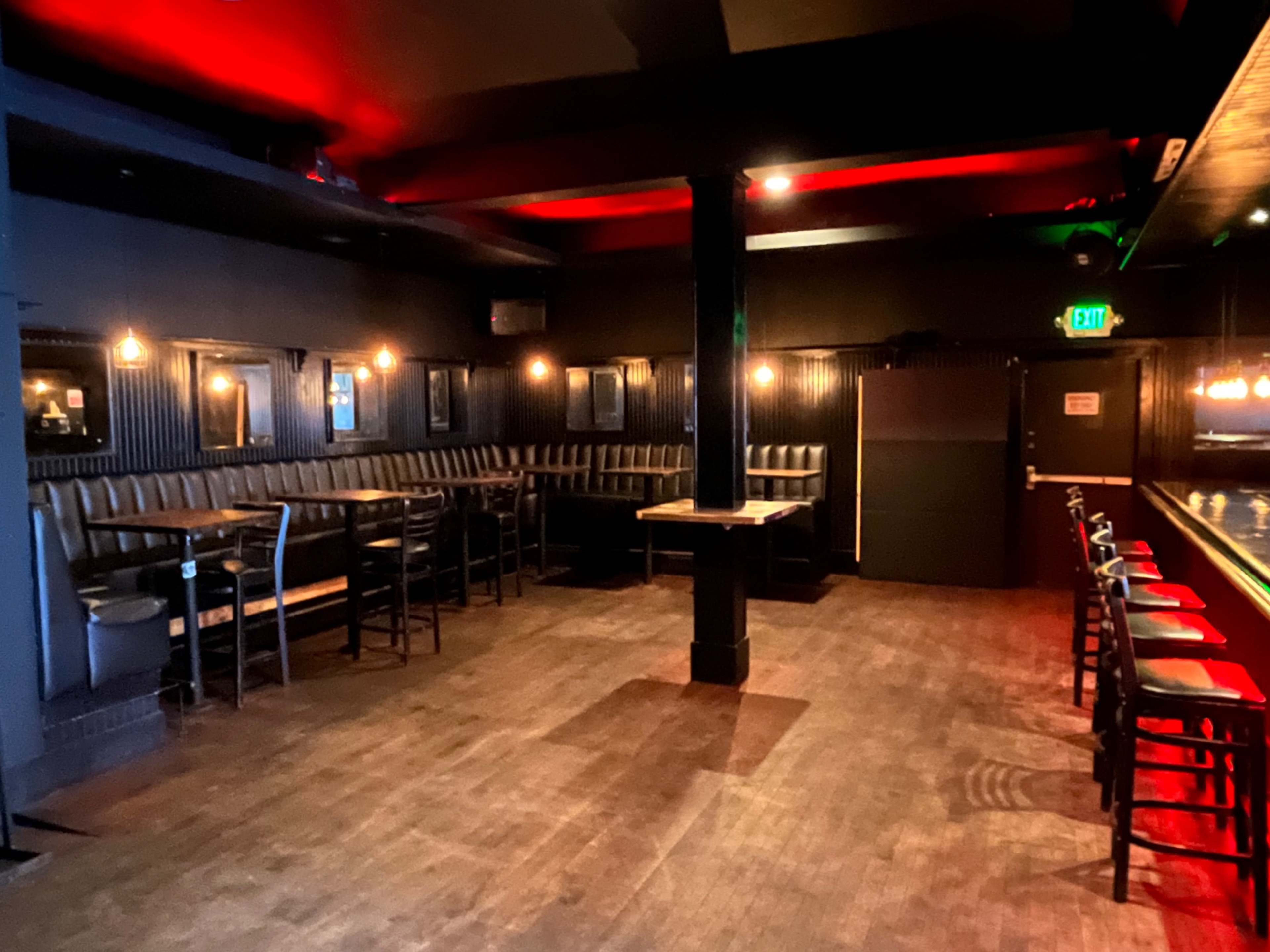A dimly lit bar interior featuring a row of black leather benches and high-top tables alongside a polished bar counter.