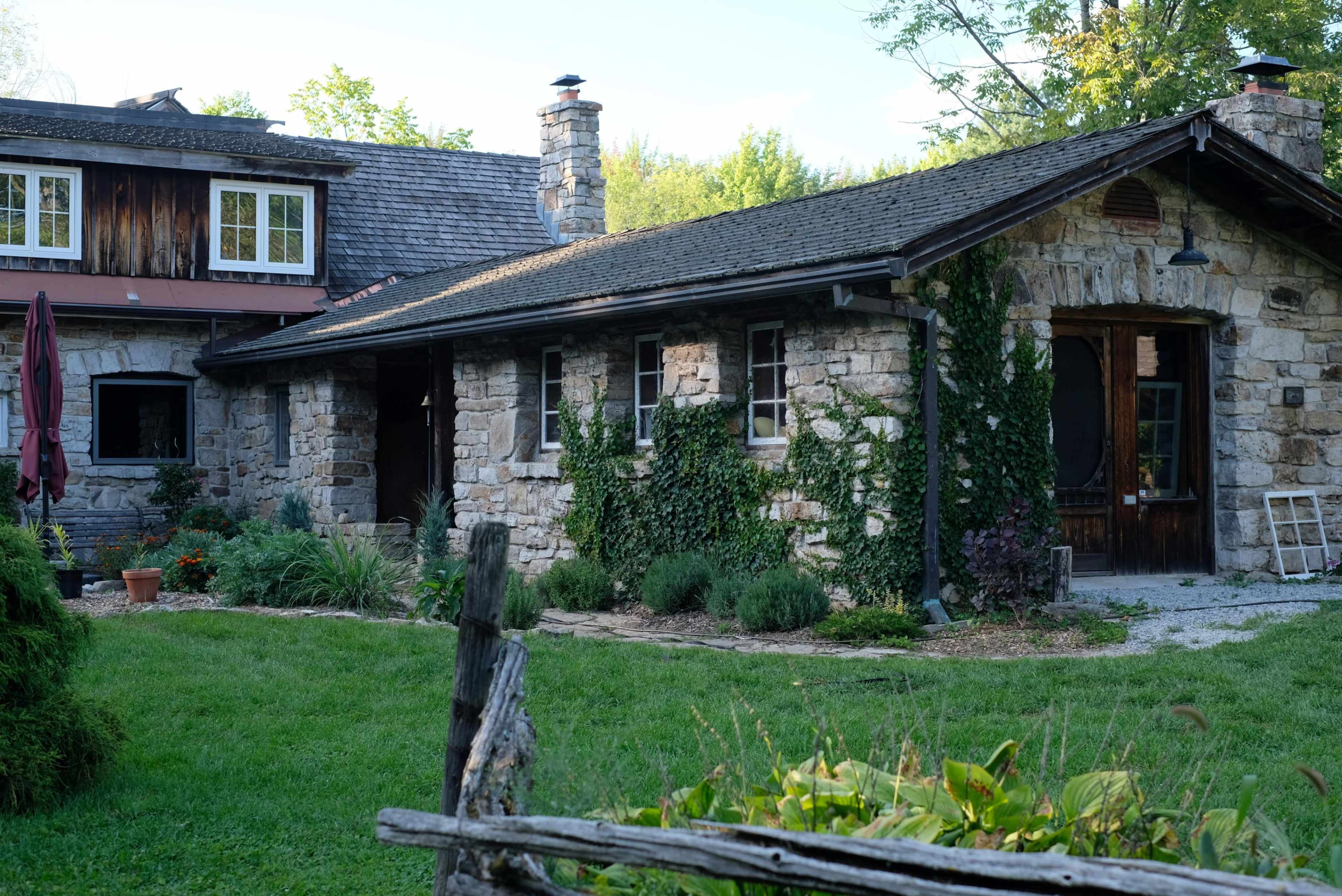 A rustic stone house surrounded by greenery, with a wooden structure adjacent to it and a garden area in front.