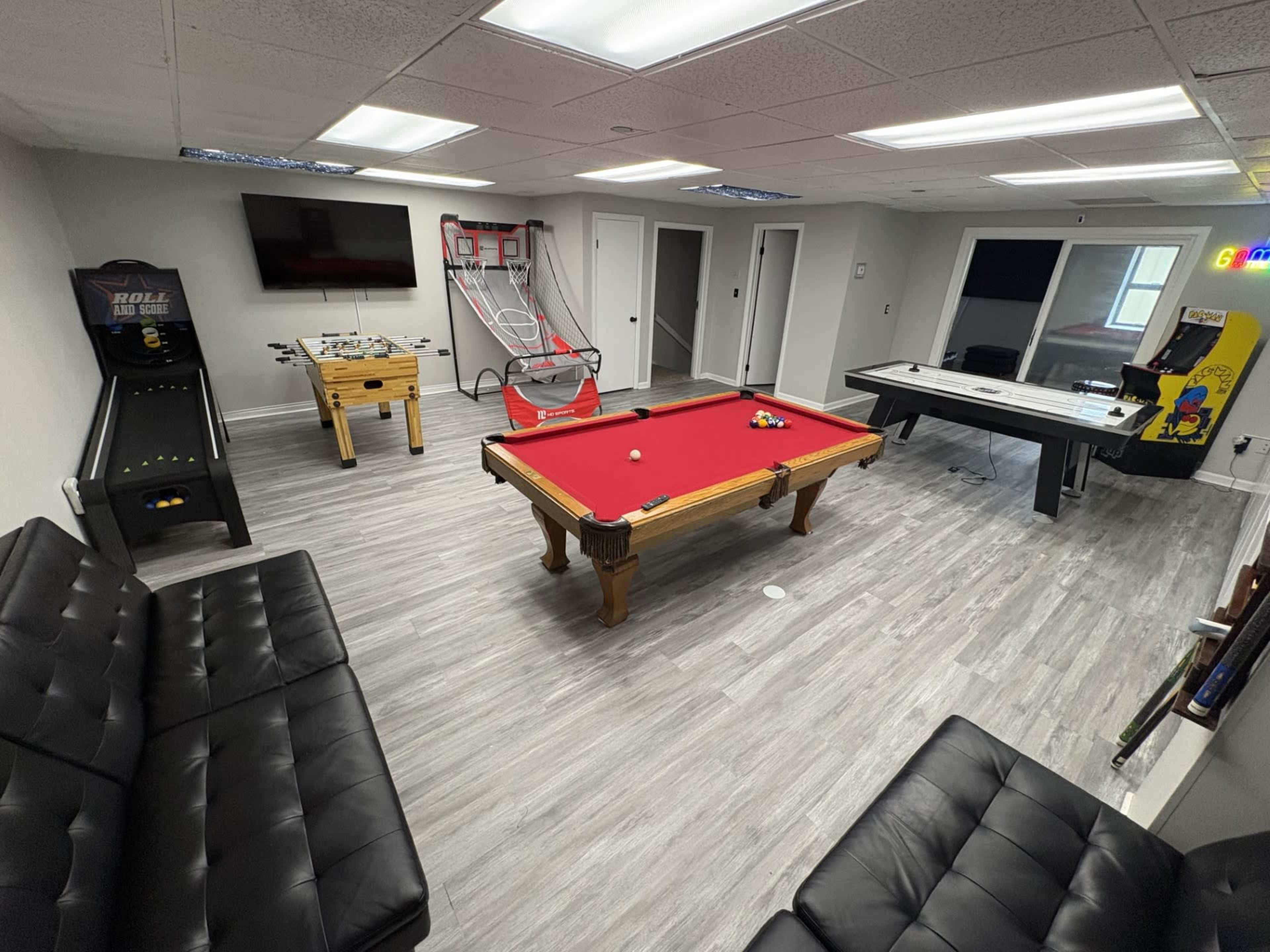 The image shows a recreational room featuring a red pool table, an arcade game machine, a foosball table, a basketball shooting game, and a table hockey setup, all surrounded by gray flooring and white walls.