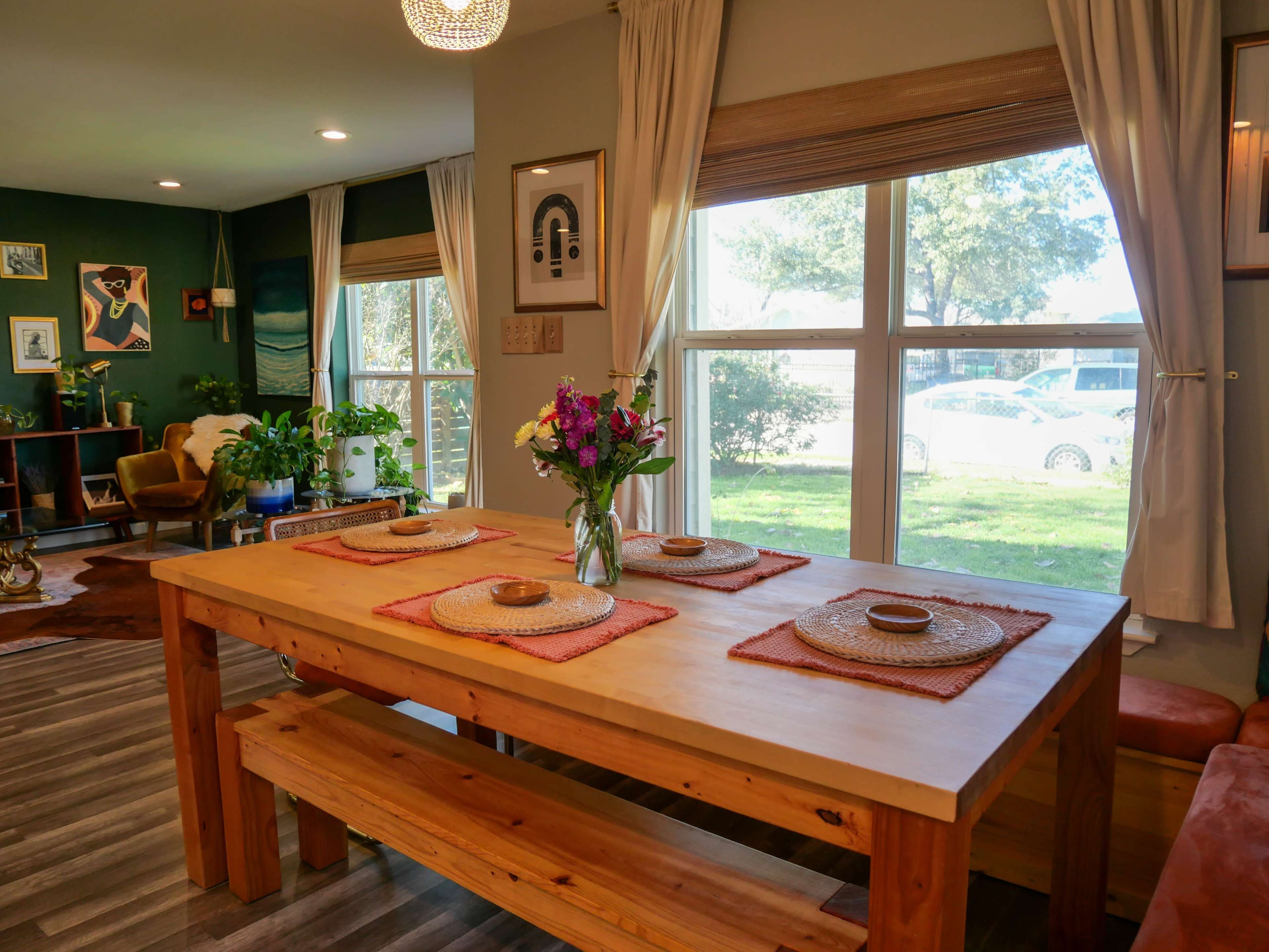 A wooden dining table with place settings and a vase of flowers is positioned near a large window overlooking a garden.