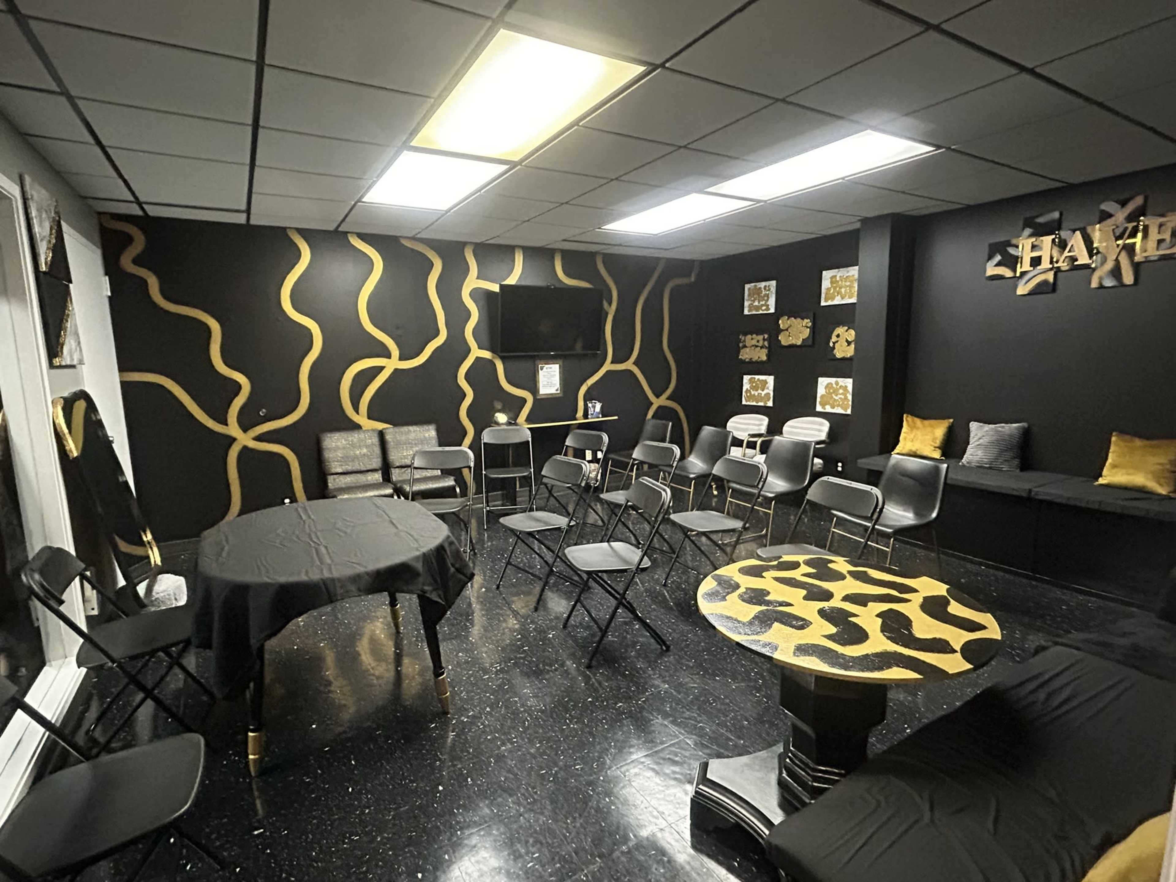The room features black walls with gold abstract designs, a central table with black cloth, folding chairs arranged around it, and a television on one wall.