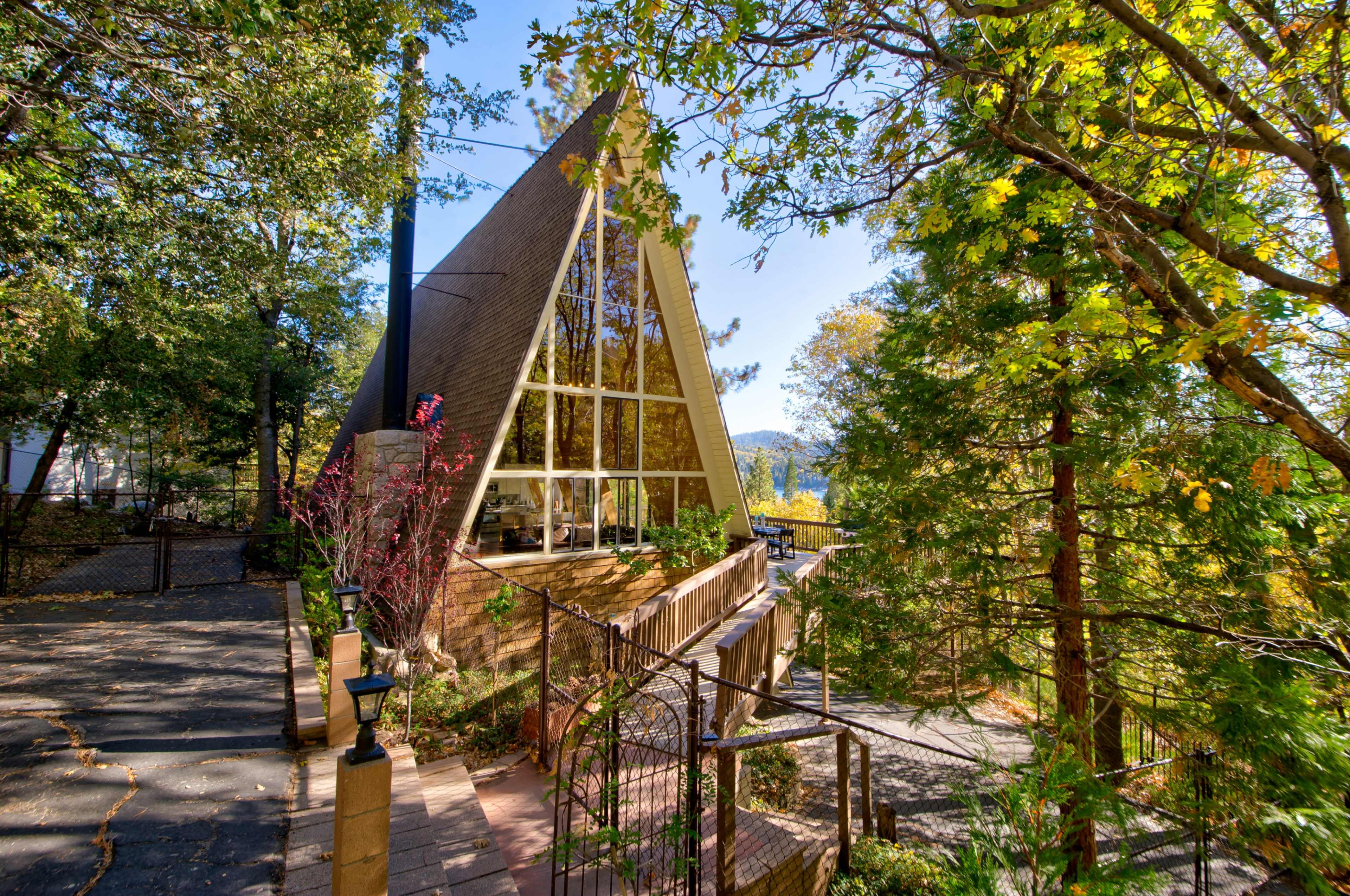 A modern A-frame house with large windows, surrounded by trees and a pathway leading to its entrance.