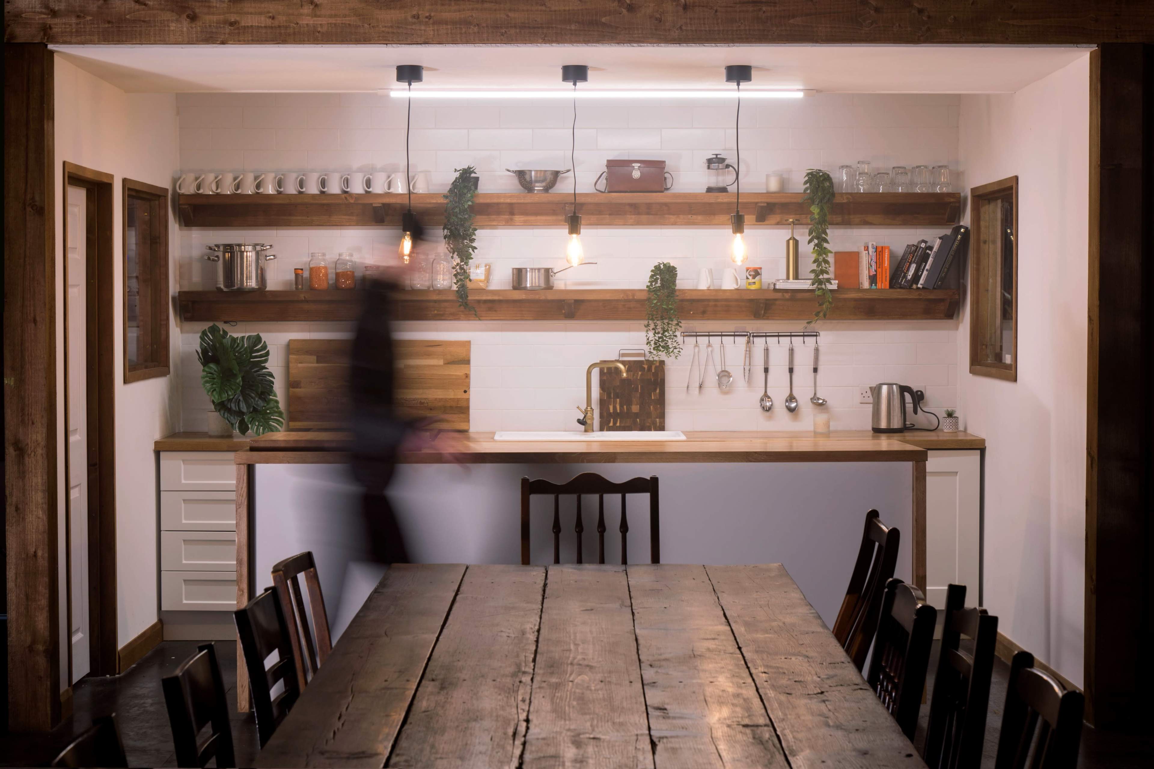A wooden table sits in the foreground of a modern kitchen featuring open shelves with various kitchenware and plants, illuminated by pendant lights.