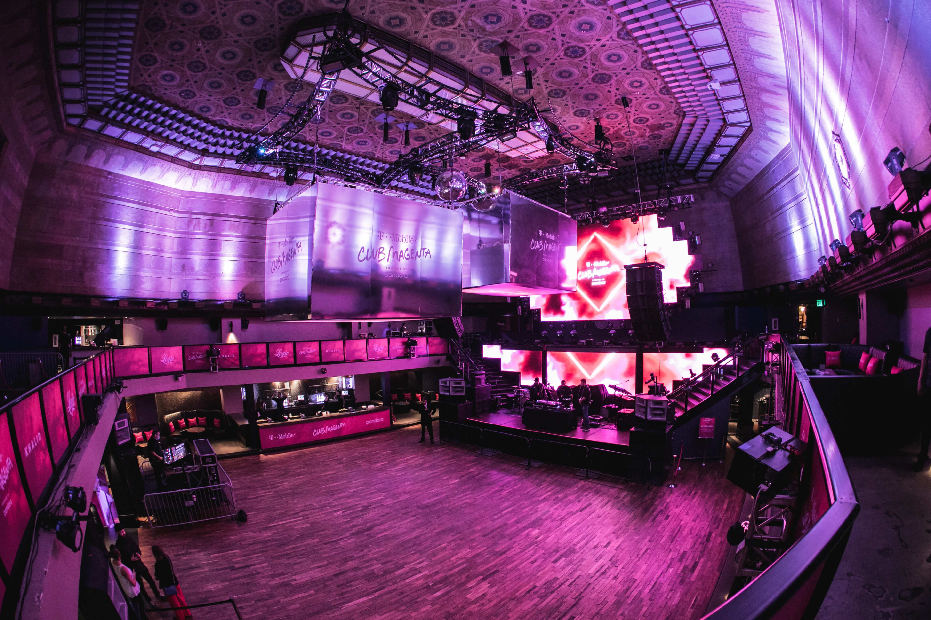 A music venue with a wide, empty floor and illuminated walls featuring large screens and colorful lighting.