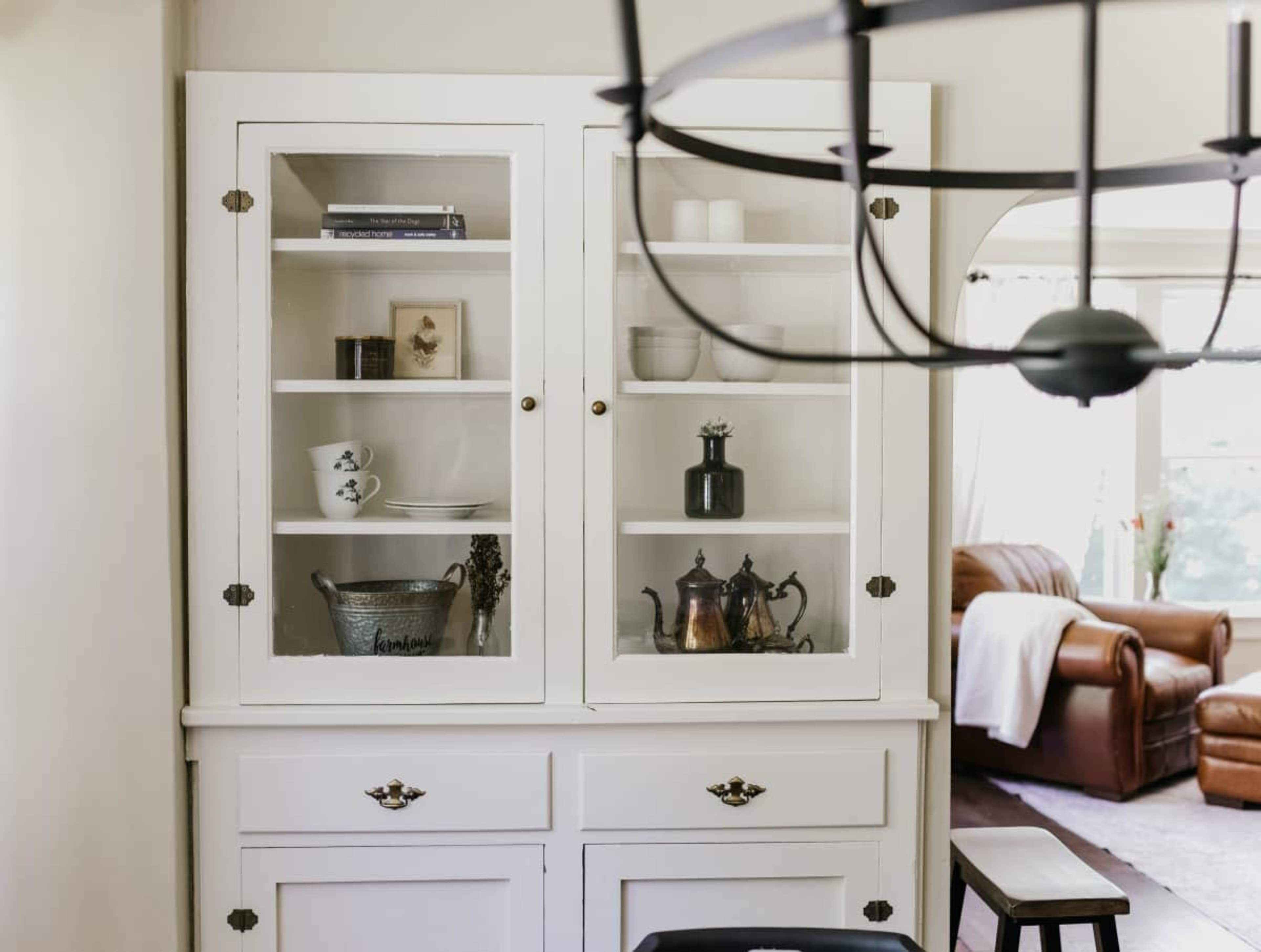 A white wooden cabinet with glass doors displays dishes and decorative items, while a leather sofa is visible in the background.