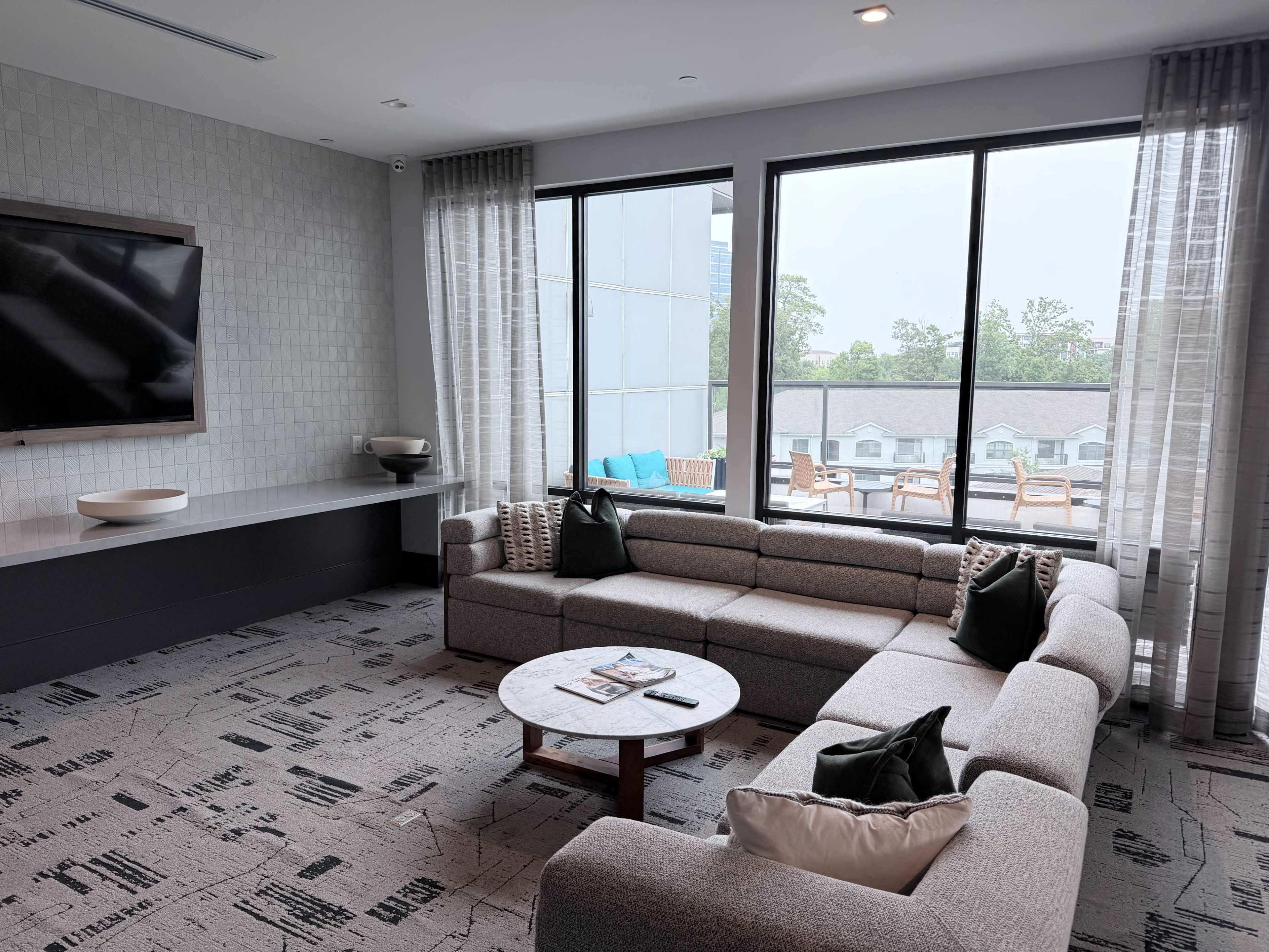 A modern lounge area features a gray sectional sofa and a round coffee table, with large windows overlooking an outdoor patio.