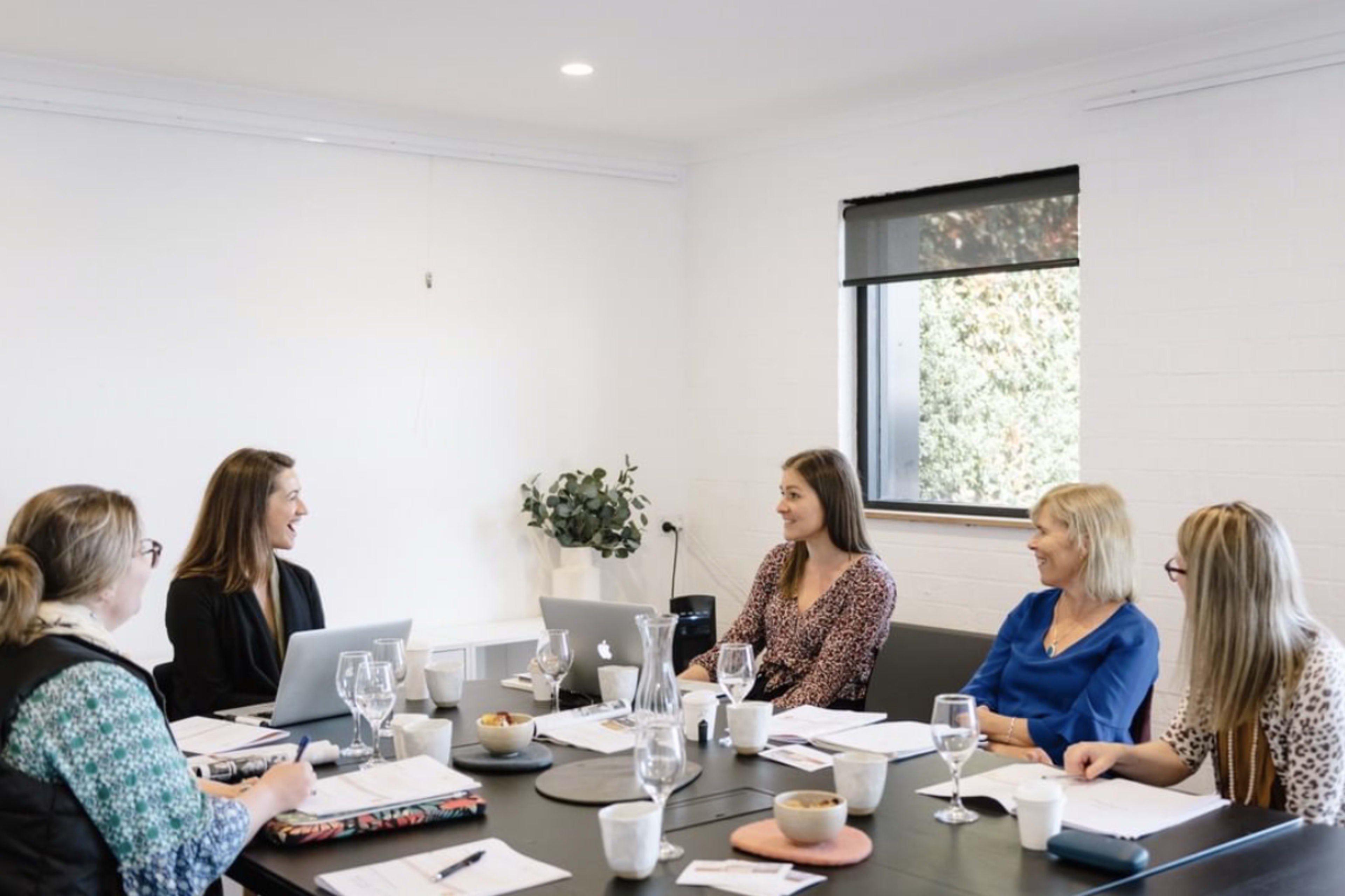A group of five women are seated around a conference table, engaged in a discussion with laptops and documents in front of them.