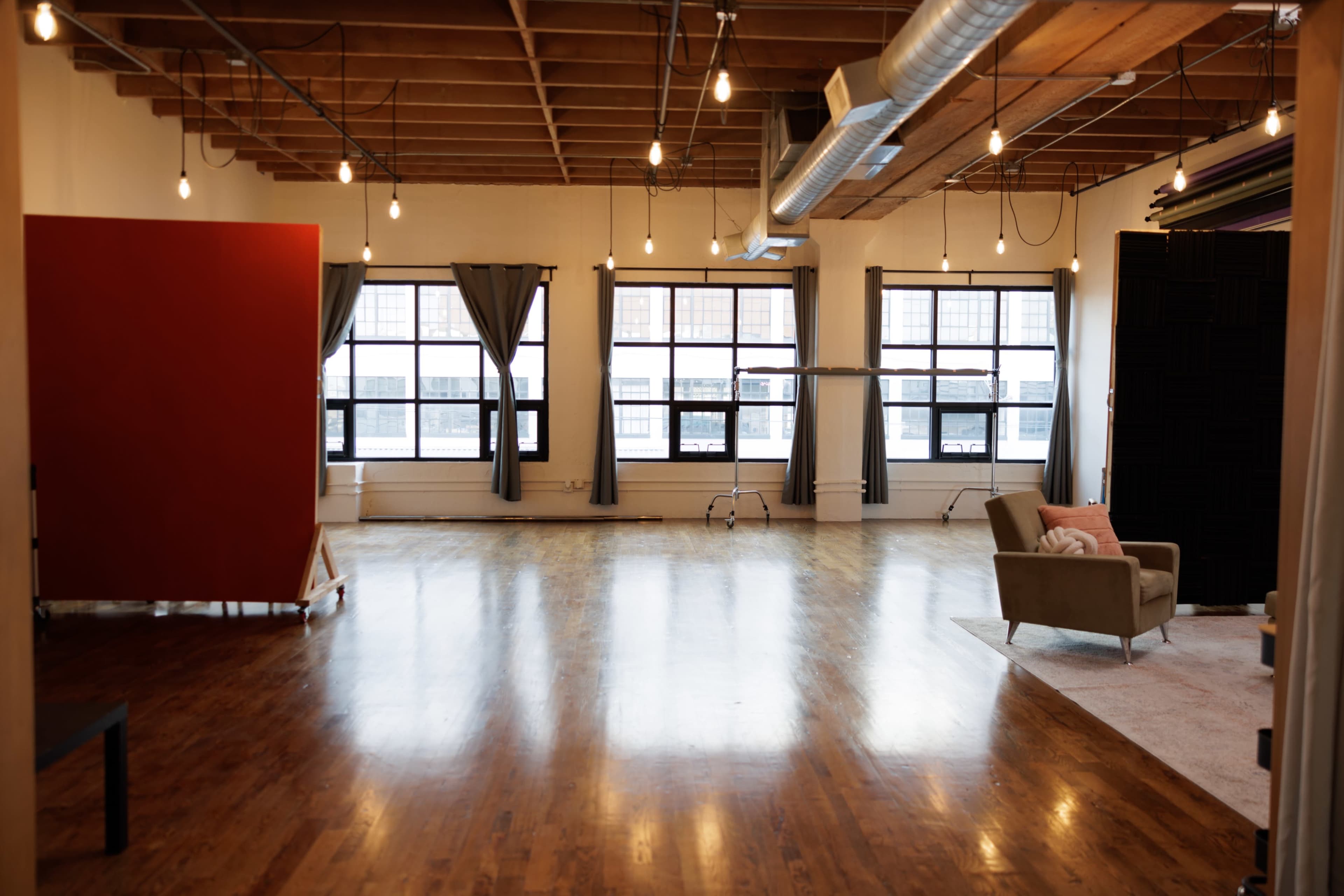 A spacious, well-lit studio features large windows, polished wooden floors, a sofa, and simple decor.