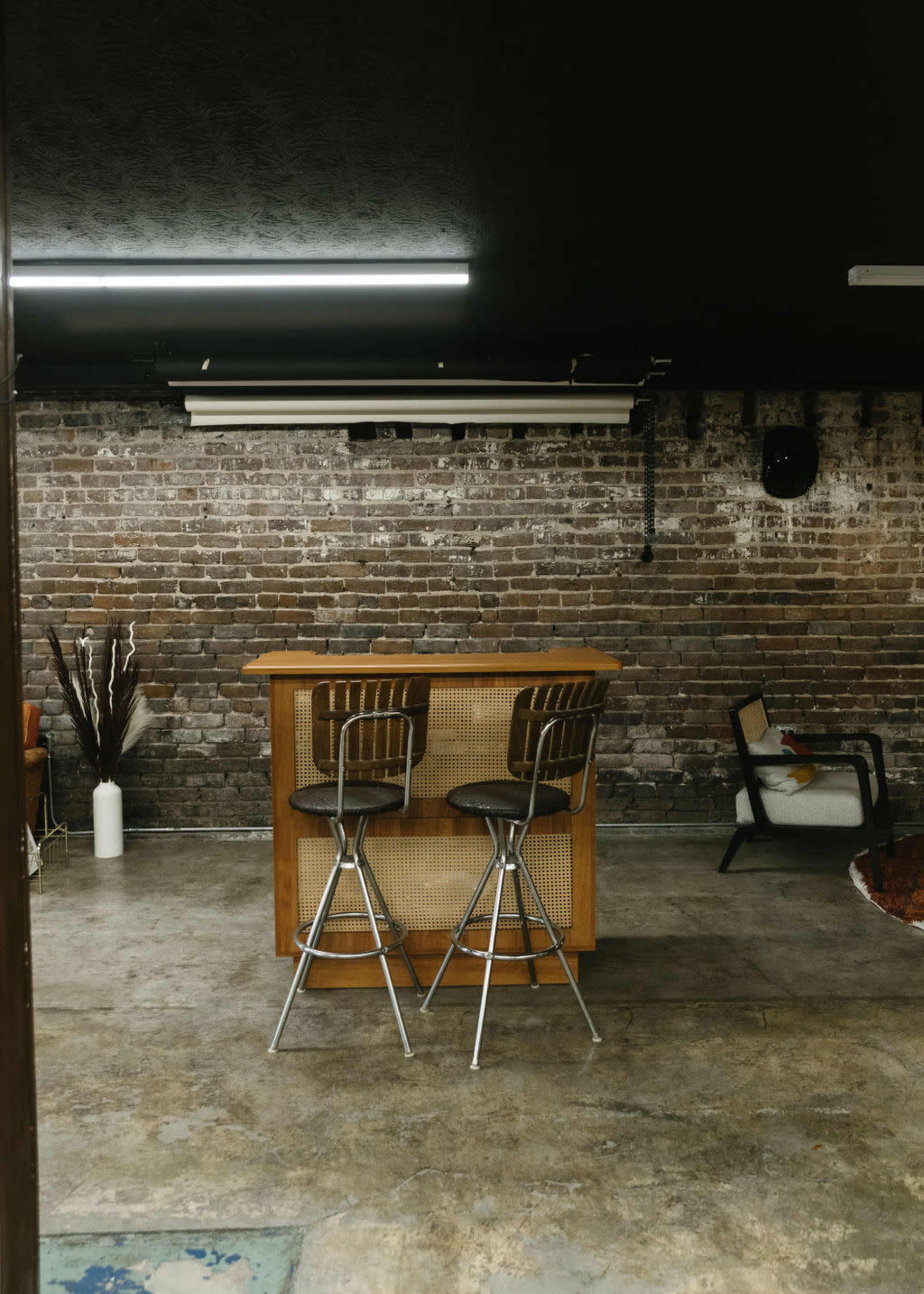 The image shows a minimalist bar area with two metal and wooden stools in front of a wooden bar against a brick wall.