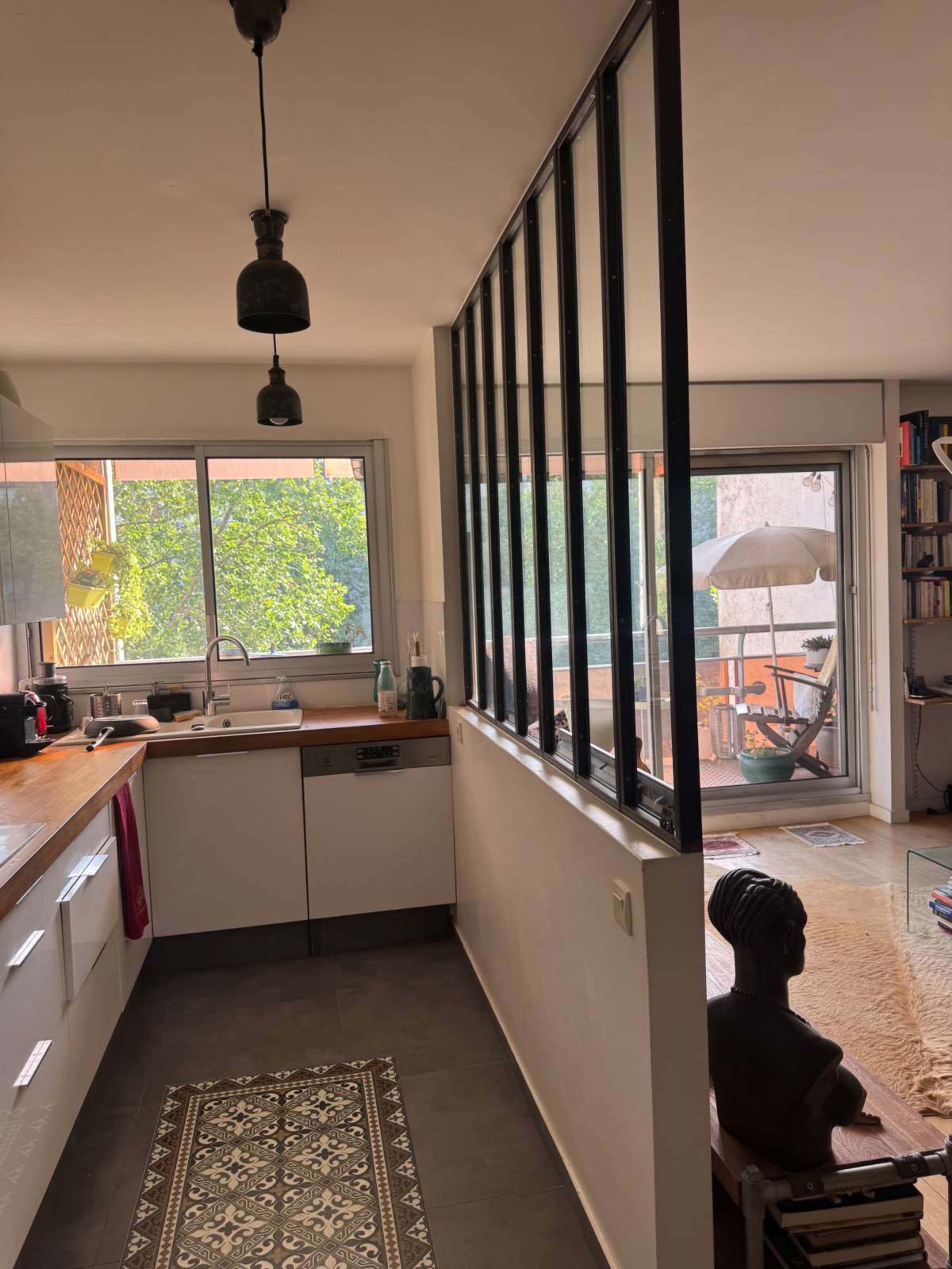 The image shows a modern kitchen with a wooden countertop, black metal partition, and a view into a bright living area containing a large window and a sculpture.