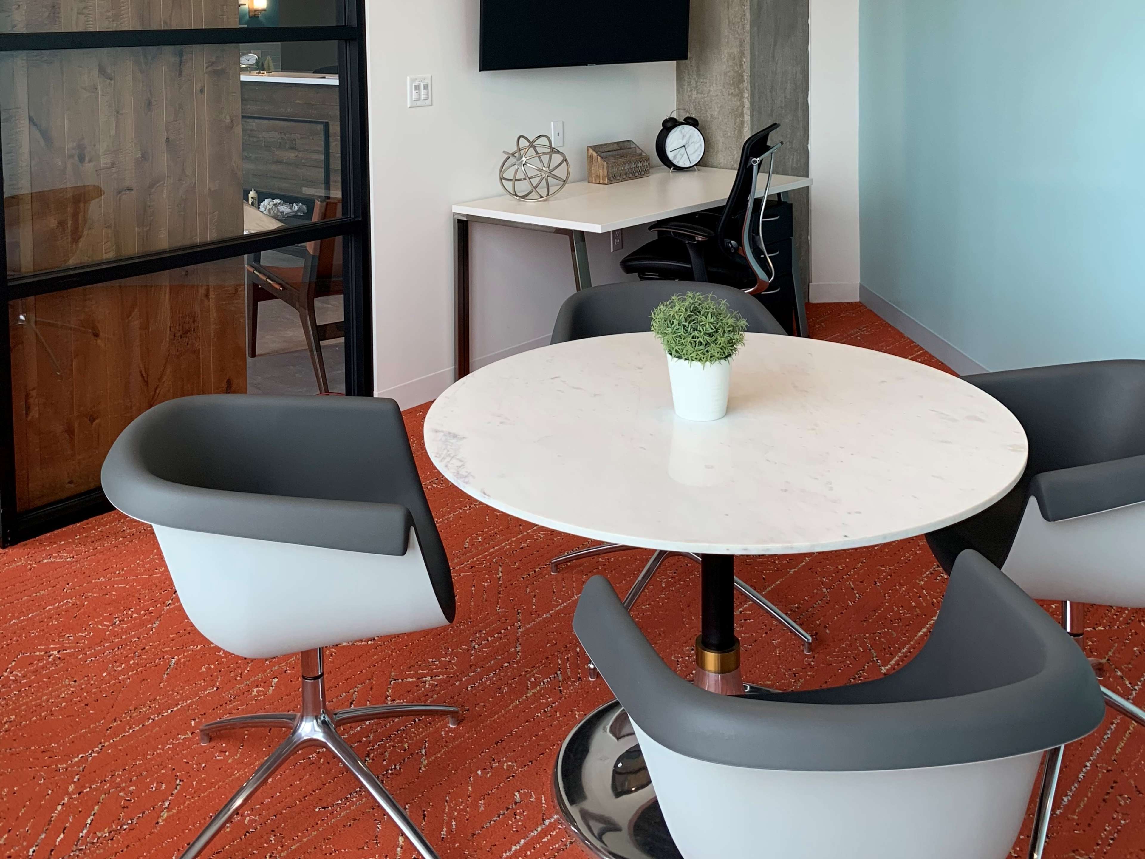 A round marble table is surrounded by four modern gray chairs in an office meeting space with orange carpet.