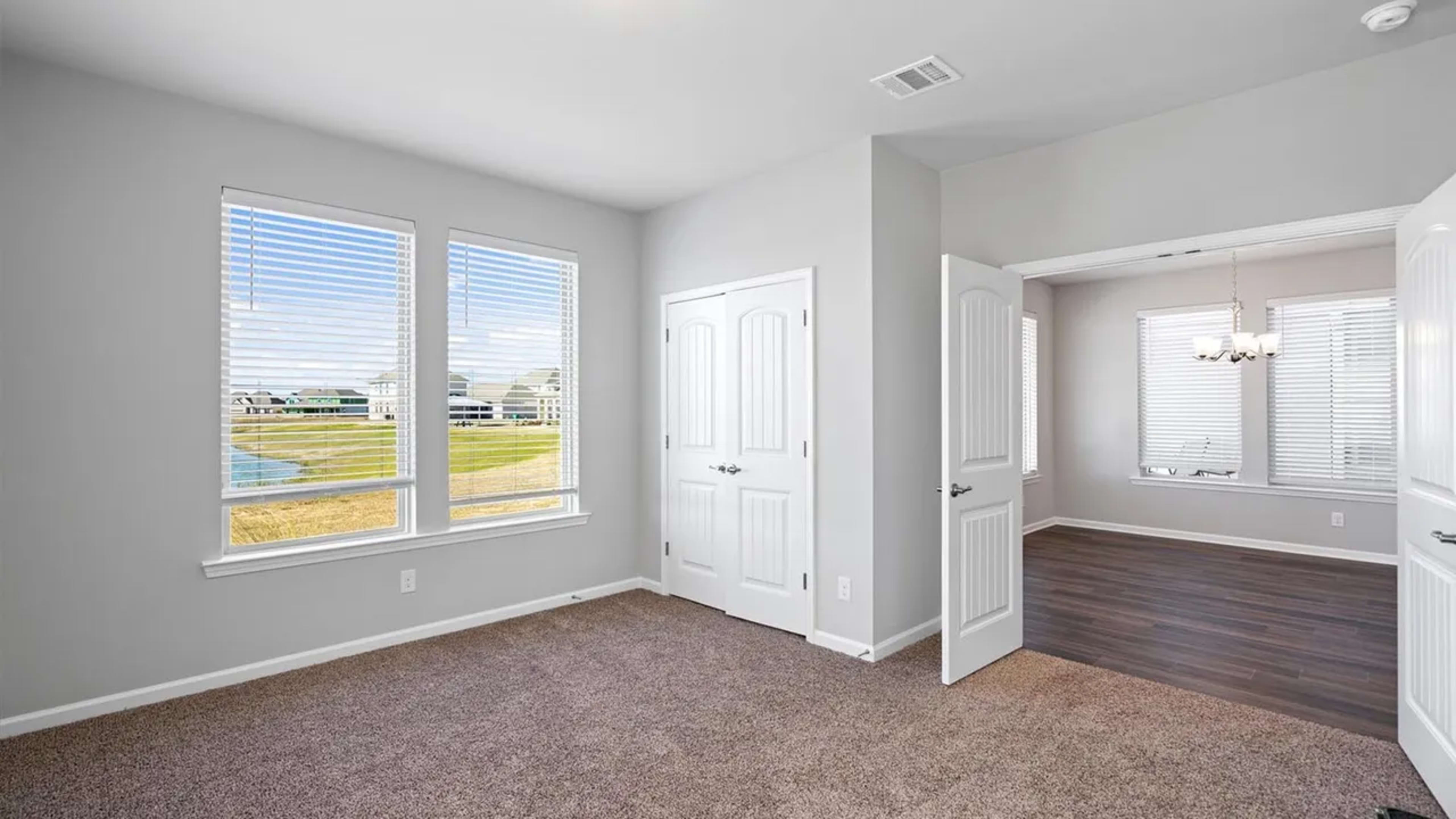 The image shows a corner of a room with two windows, freshly painted light gray walls, and beige carpet, leading into an adjoining room with a window and a chandelier.