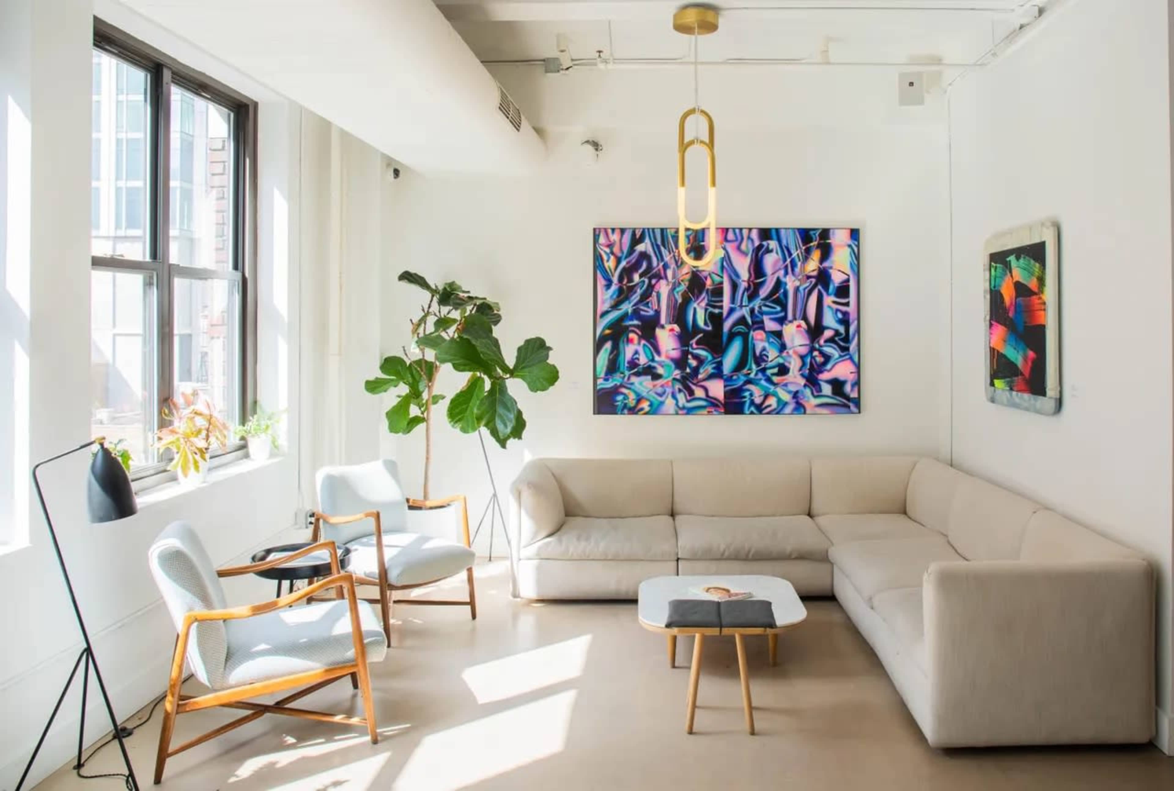 The image shows a modern living room with a light-colored sofa, wooden chairs, a coffee table, large windows, and abstract artworks on the walls.