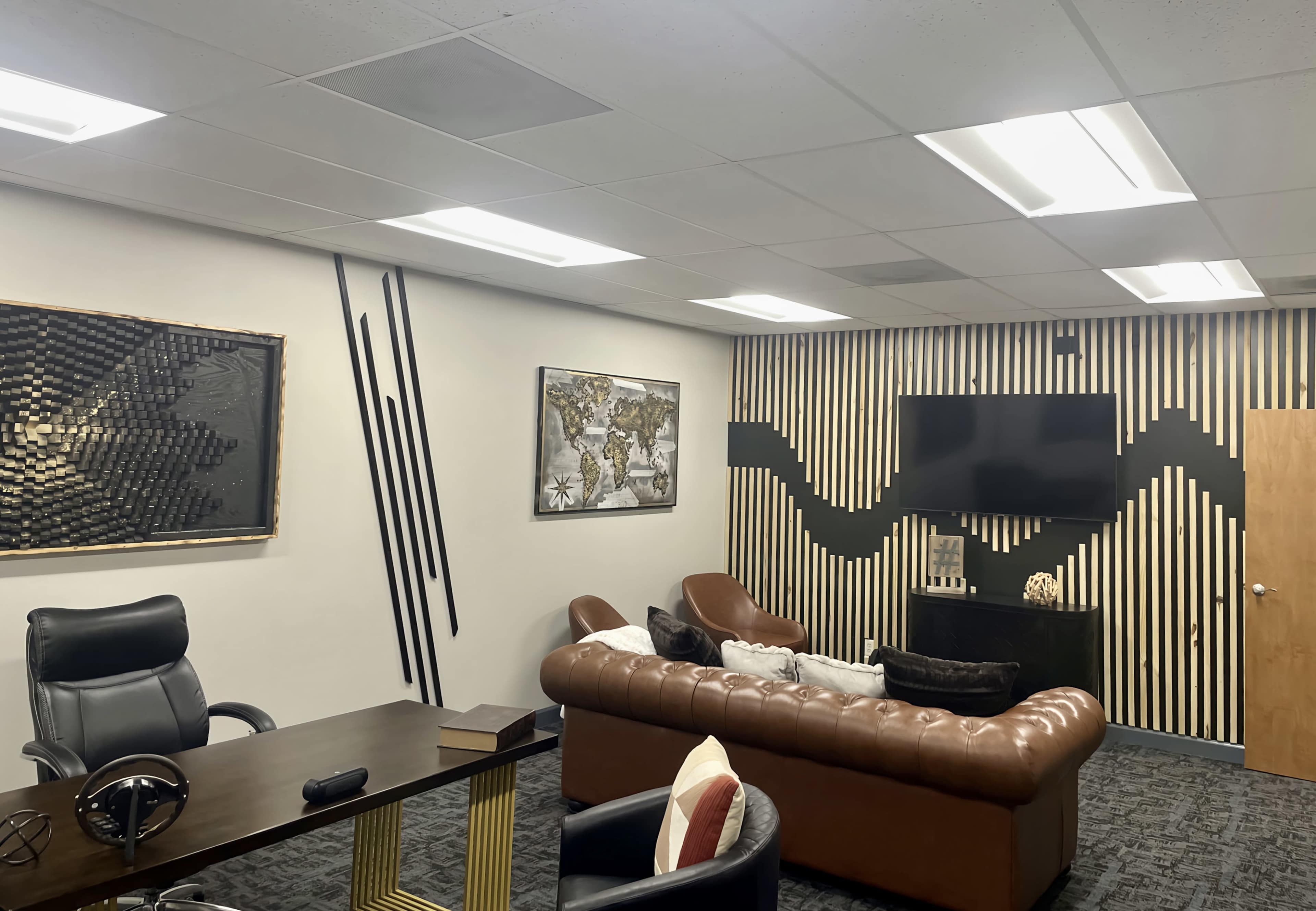 A modern office lounge area with a brown leather sofa, a wooden desk, a wall-mounted television, and decorative wall art featuring a world map and geometric designs.