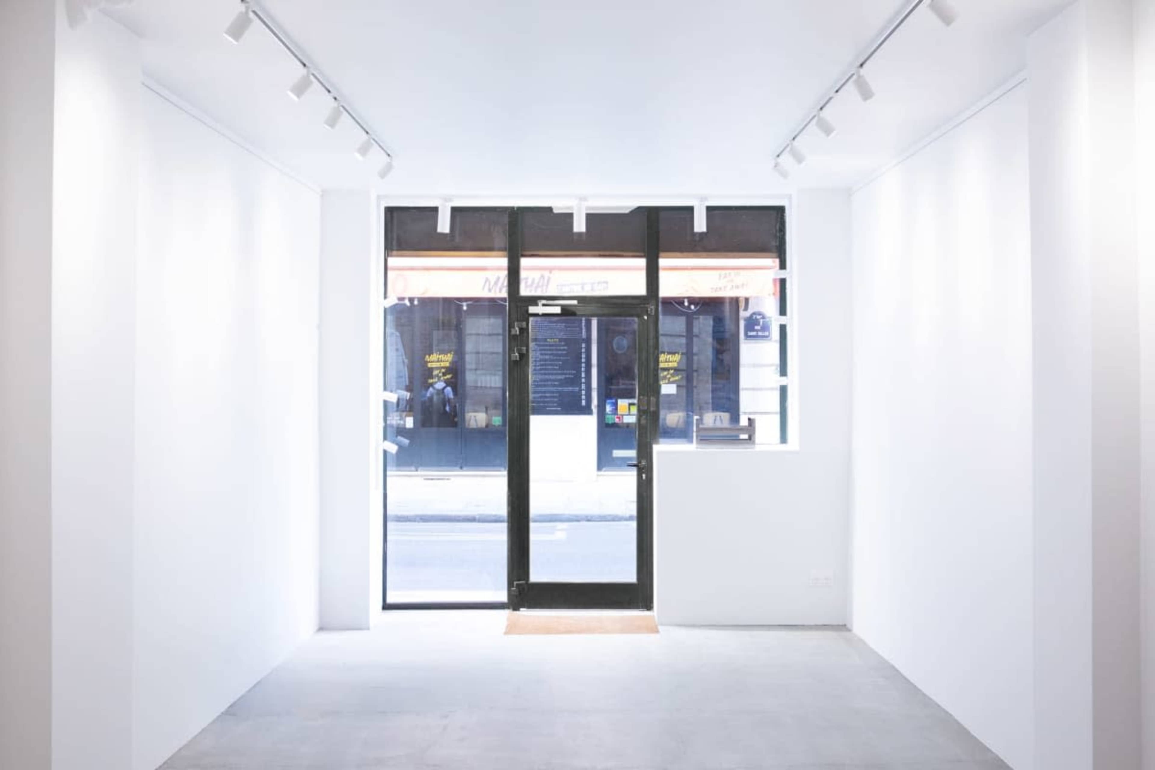 The image shows a minimalist white interior of a storefront with large windows and a black glass door, revealing the street outside.