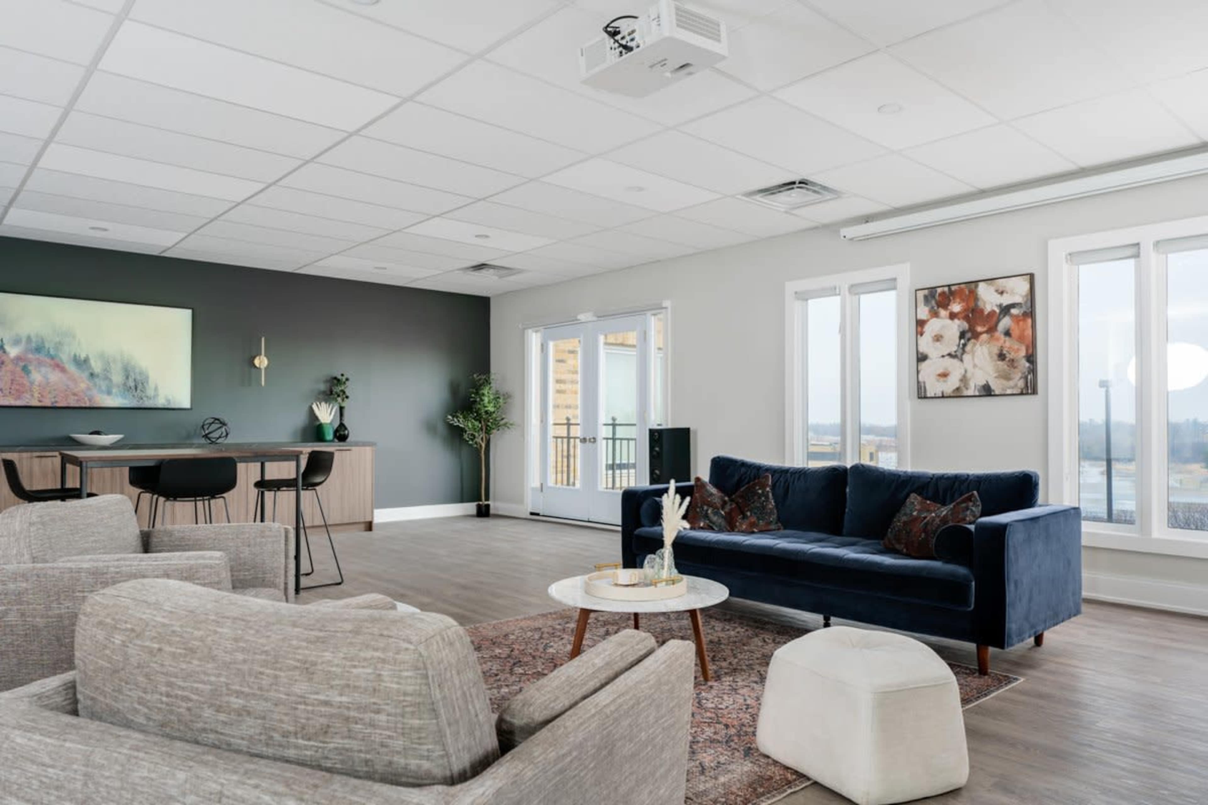 The image shows a modern living room with a navy blue sofa, light gray armchairs, a round ottoman, and large windows overlooking a view.