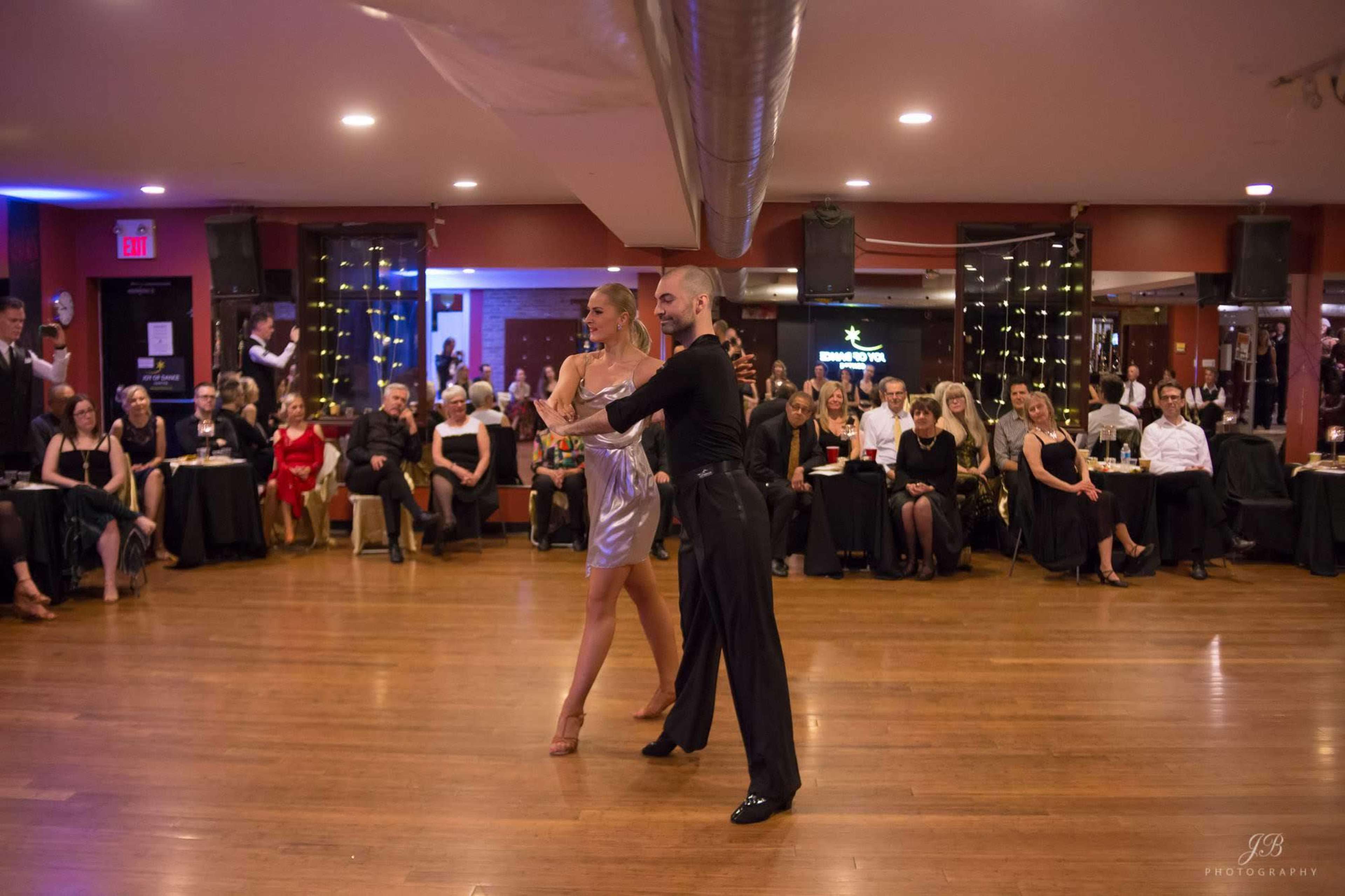 A couple performs a dance on a wooden floor in front of an audience seated at tables in a dimly lit ballroom.
