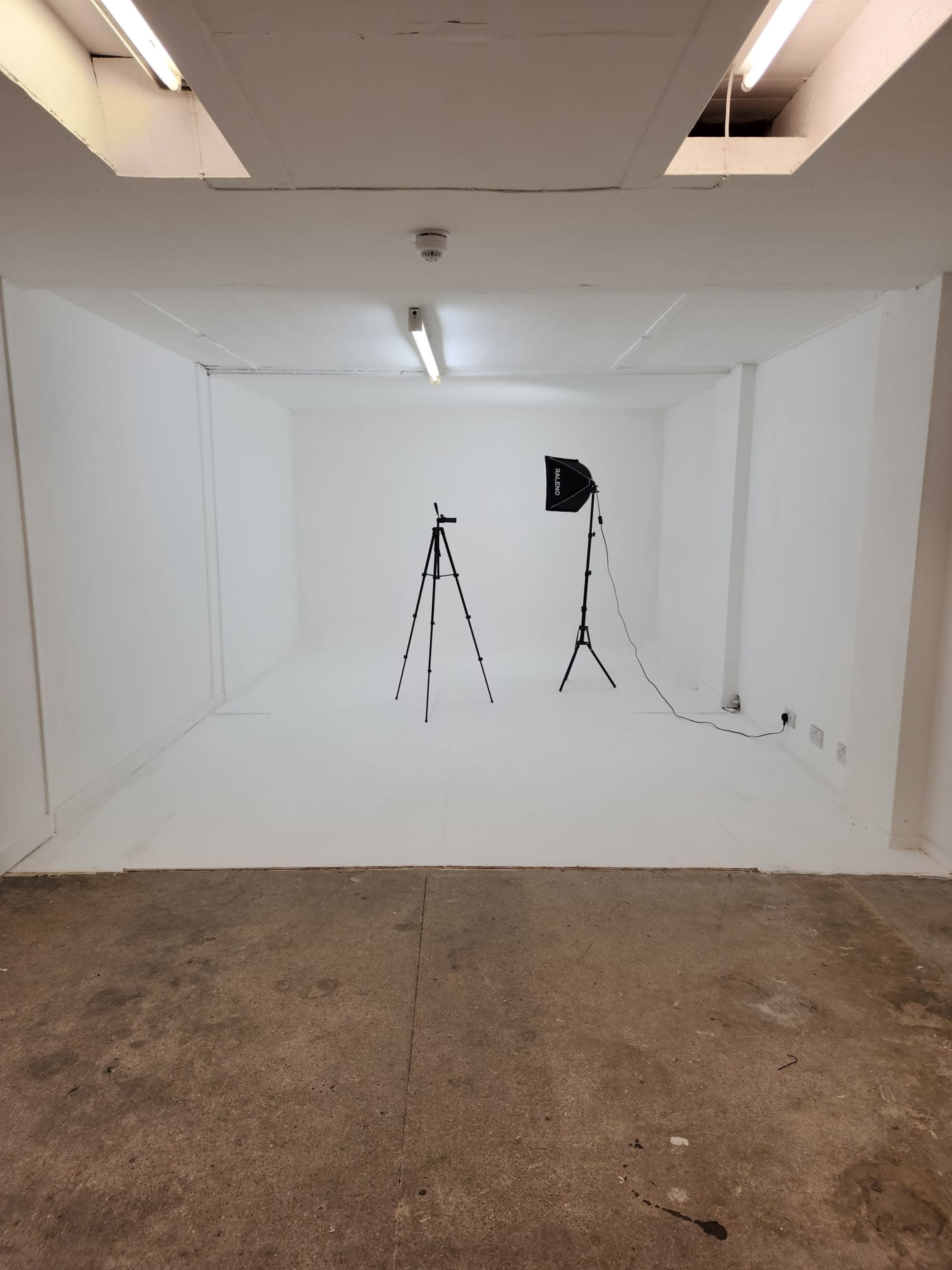 The image shows a minimalist white studio space with a concrete floor, a camera tripod, and a lighting setup.