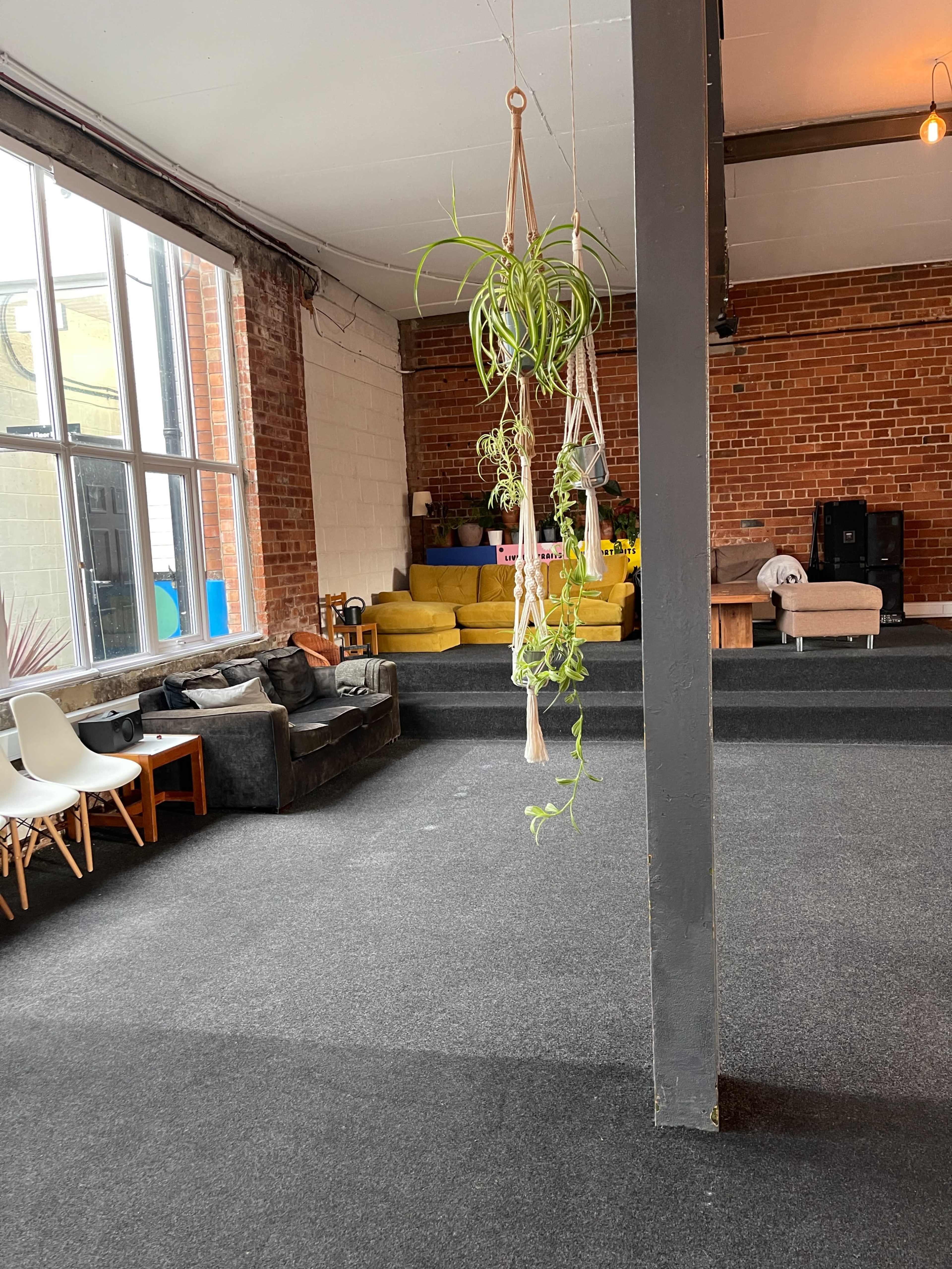A spacious indoor area with a carpeted floor, featuring a large yellow sofa and a dark-colored couch, along with hanging plants and a wall of windows.