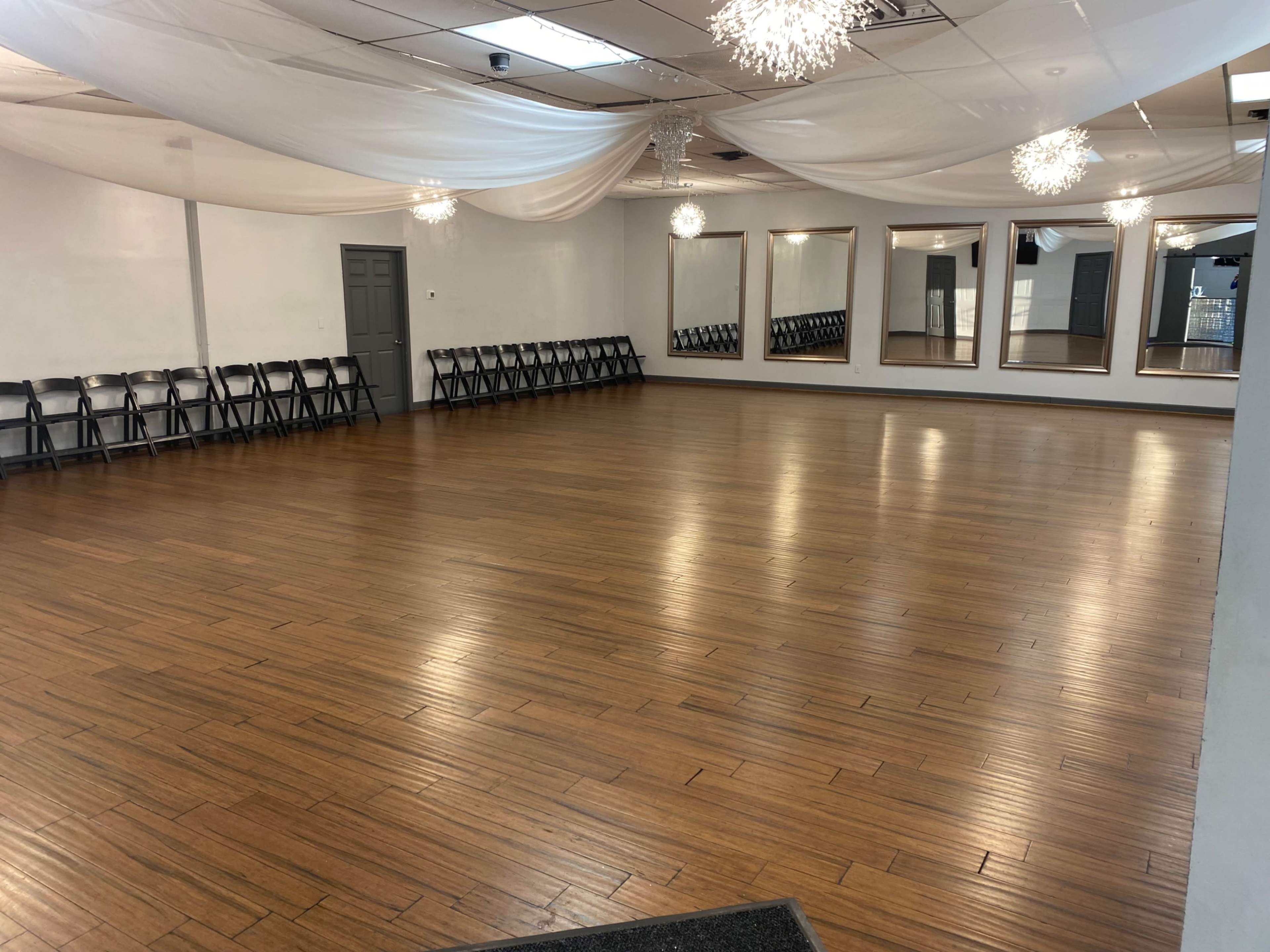 A spacious dance studio with polished wood flooring, framed mirrors on the wall, and black folding chairs arranged along the sides.