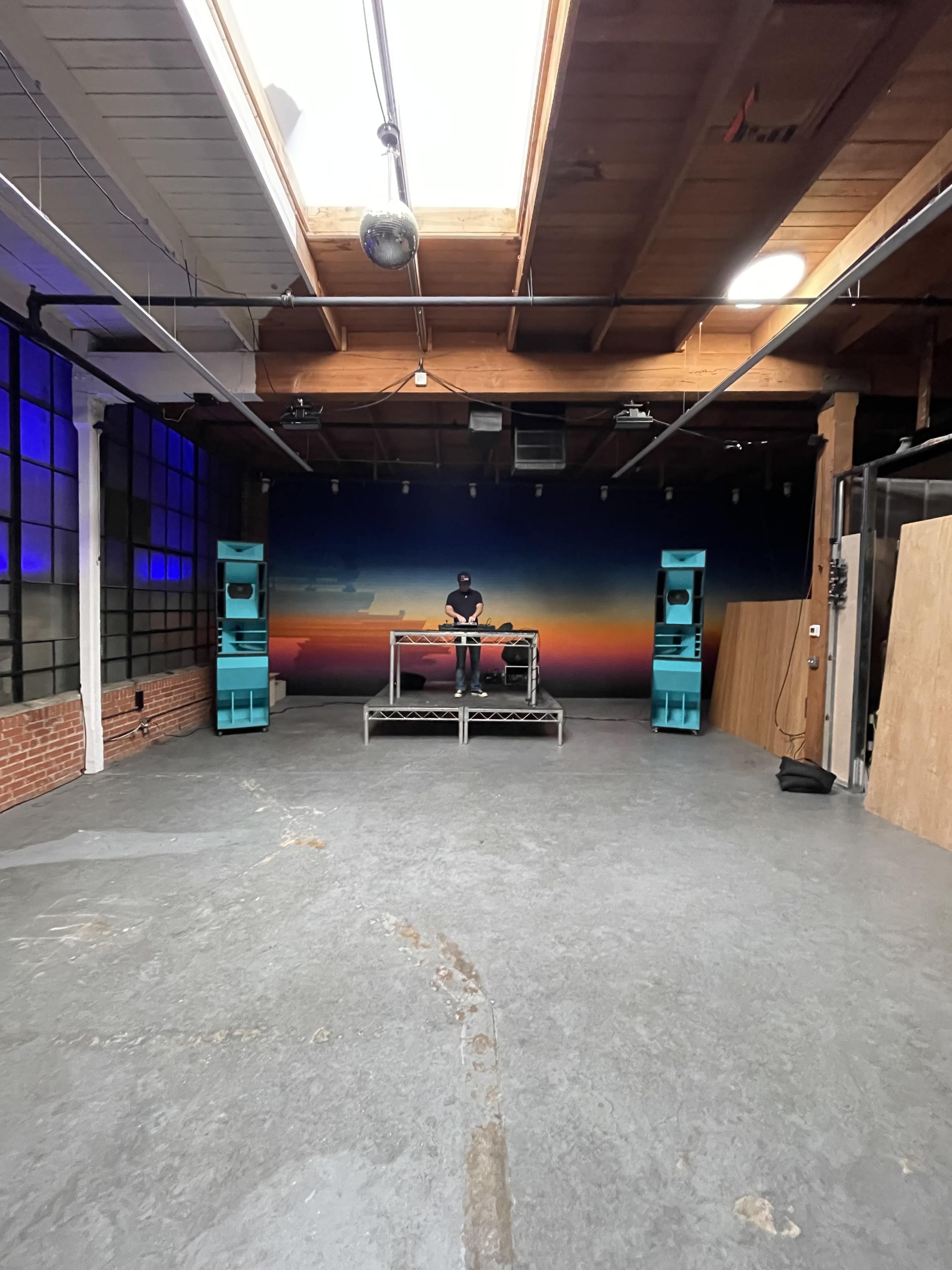 A DJ stands behind a console on a raised platform in a spacious room with large windows and a sunset mural on the wall.