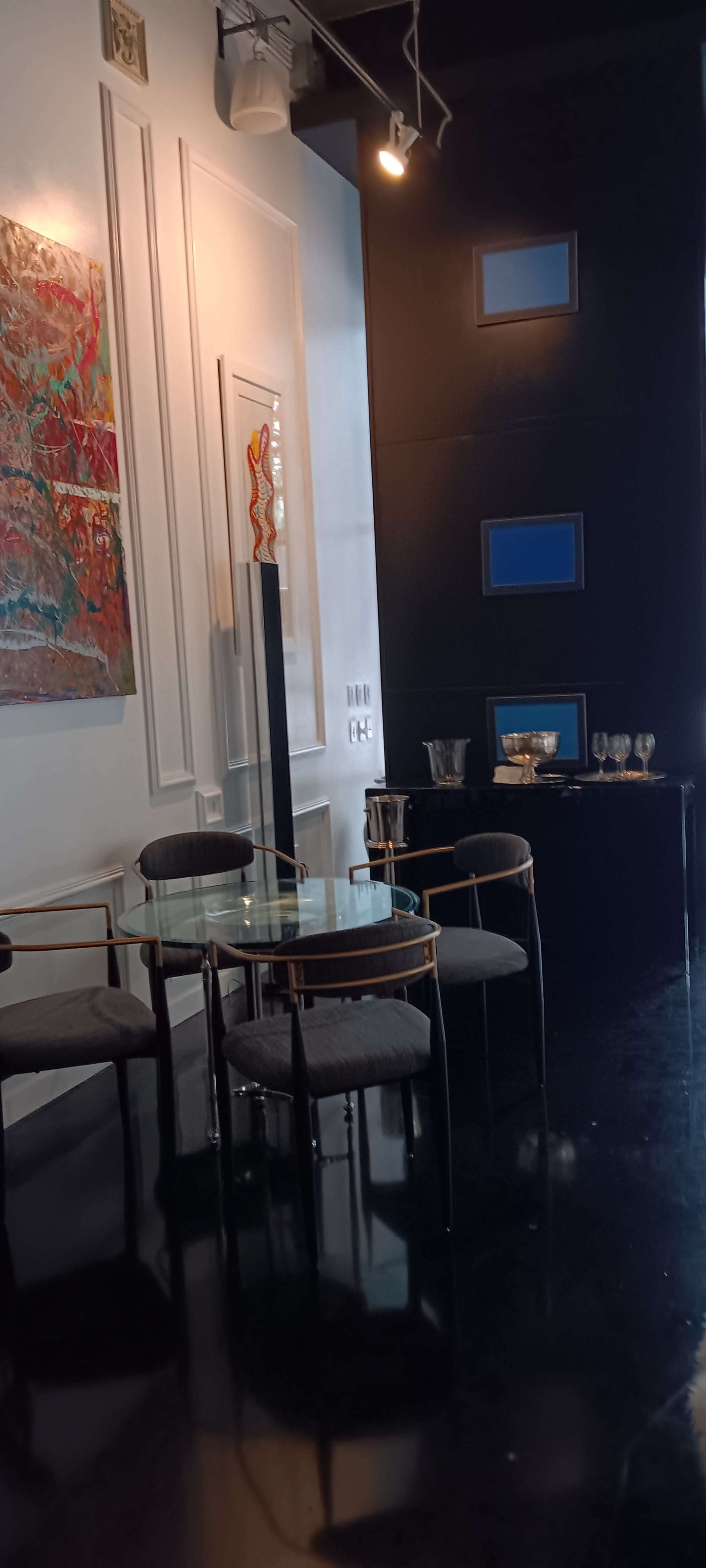 The image shows a modern interior space featuring a glass table with four upholstered chairs, a colorful artwork on the wall, and a black cabinet with glassware.