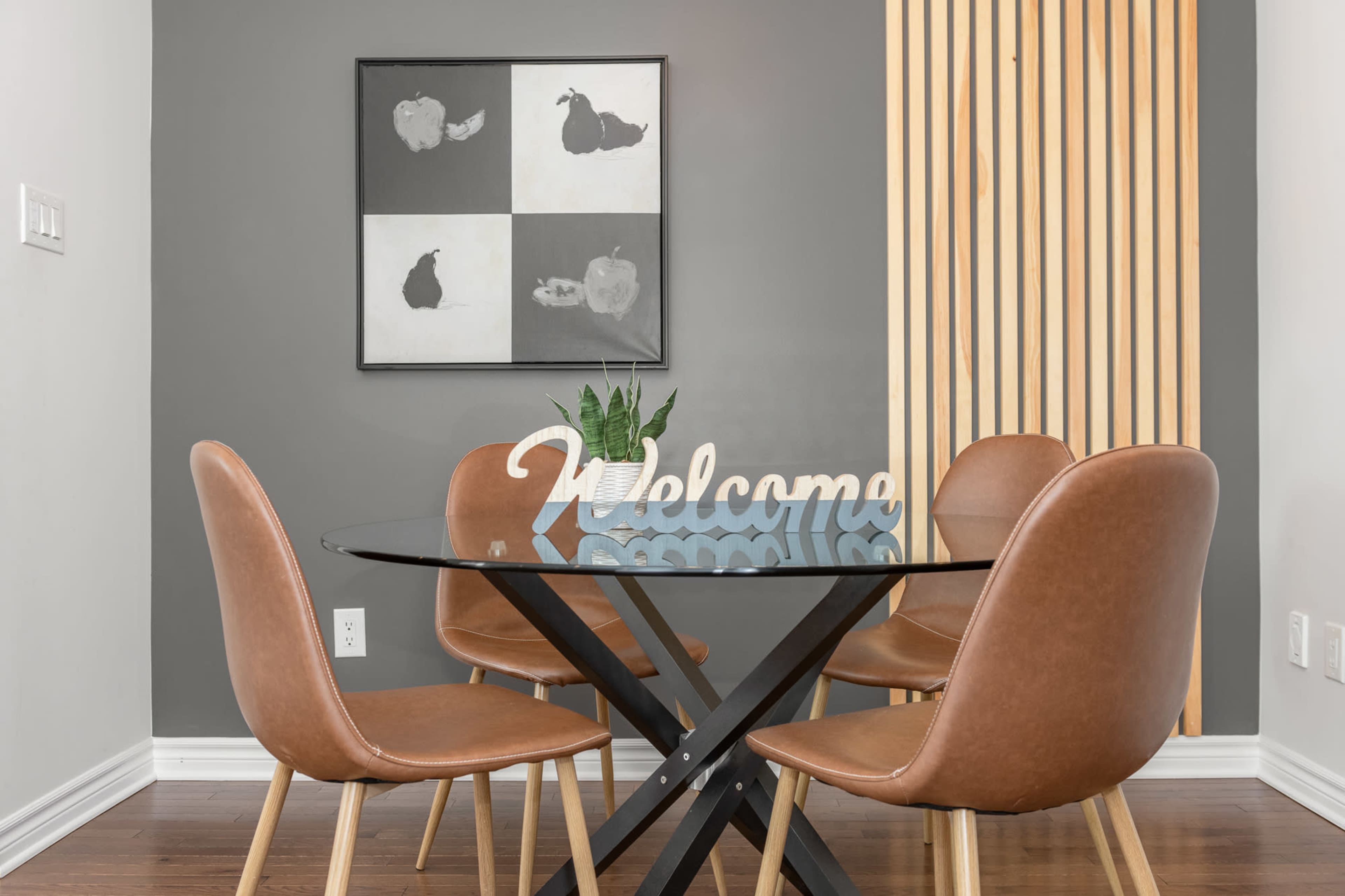 A round glass dining table with brown leather chairs is set against a gray wall featuring a black and white artwork and wooden slats.