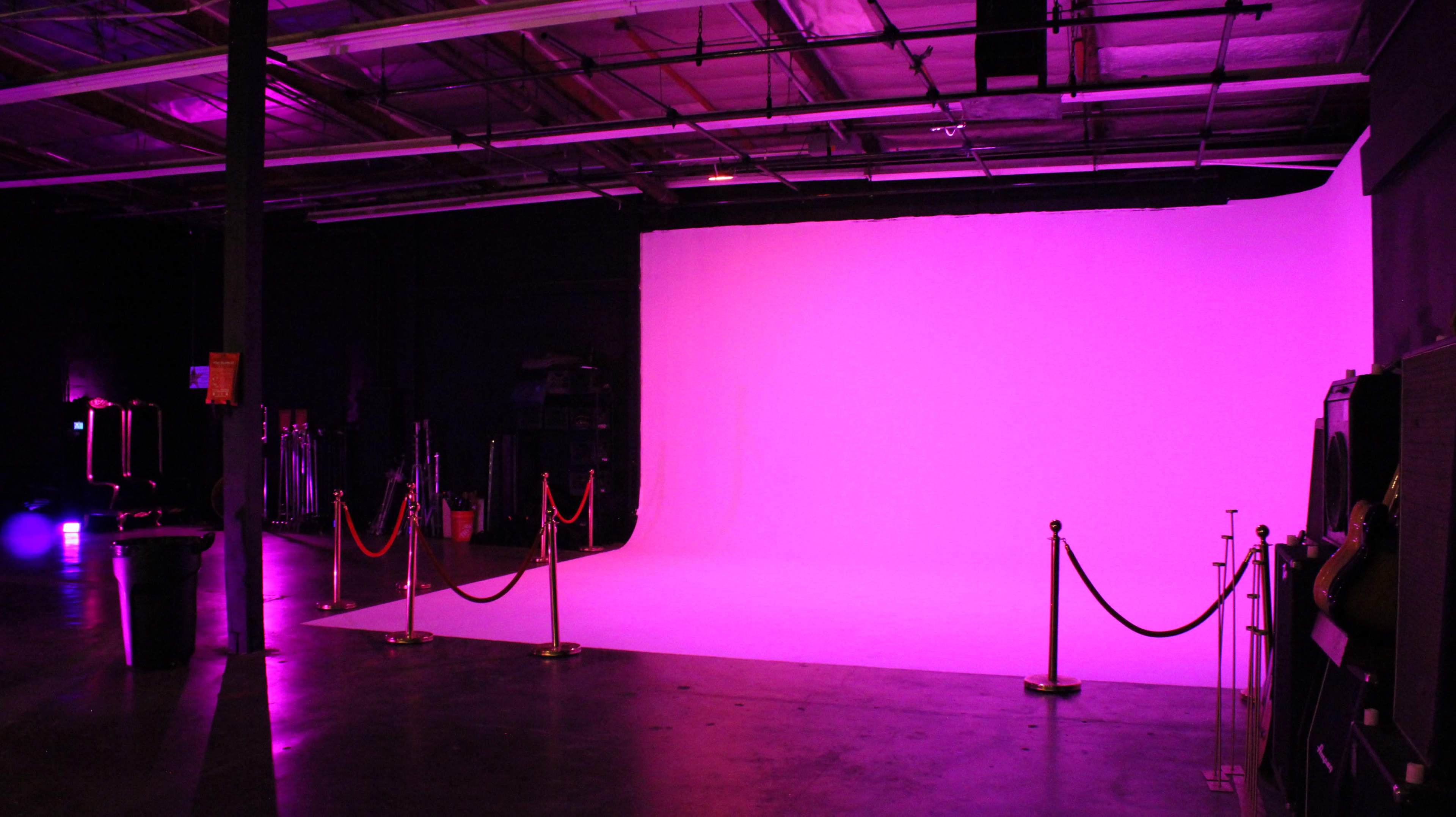 The image shows a photography studio with a large pink backdrop and illuminated floor space, surrounded by velvet ropes.