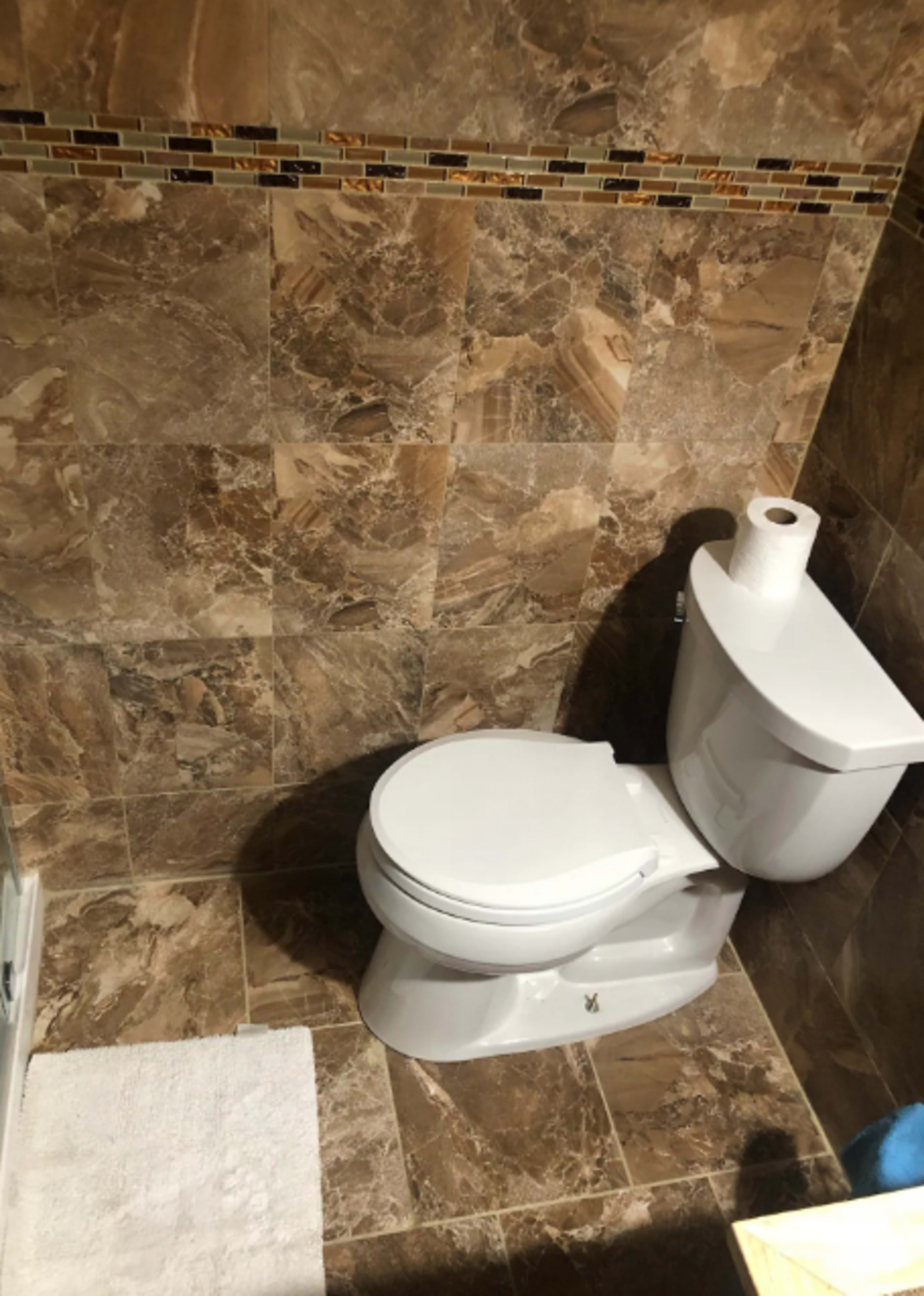 A bathroom with a white toilet positioned on a tiled floor featuring brown marble patterns and a small white rug nearby.