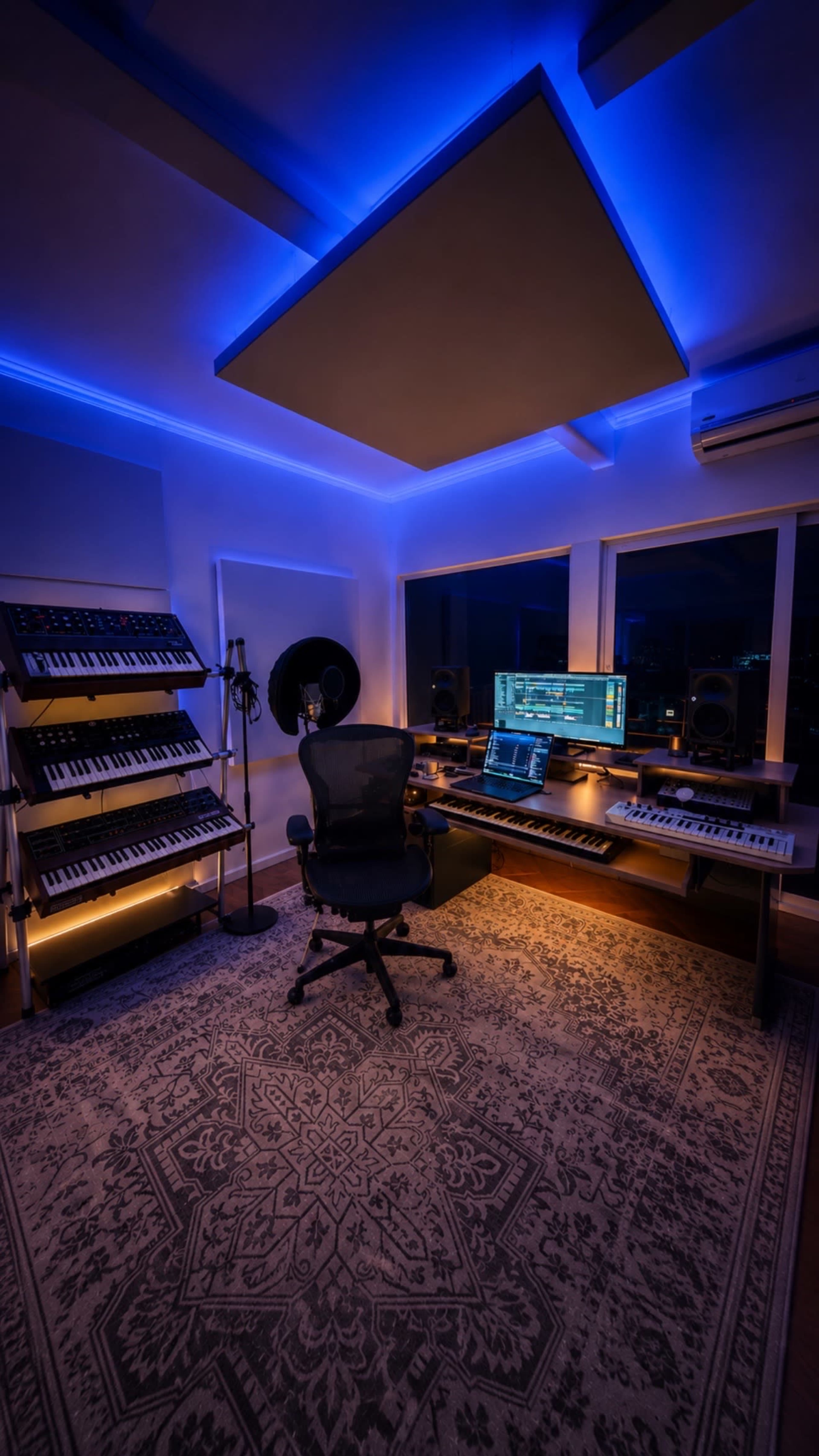 A music studio features a computer setup with audio equipment, synthesizers, and blue LED lighting illuminating the space.