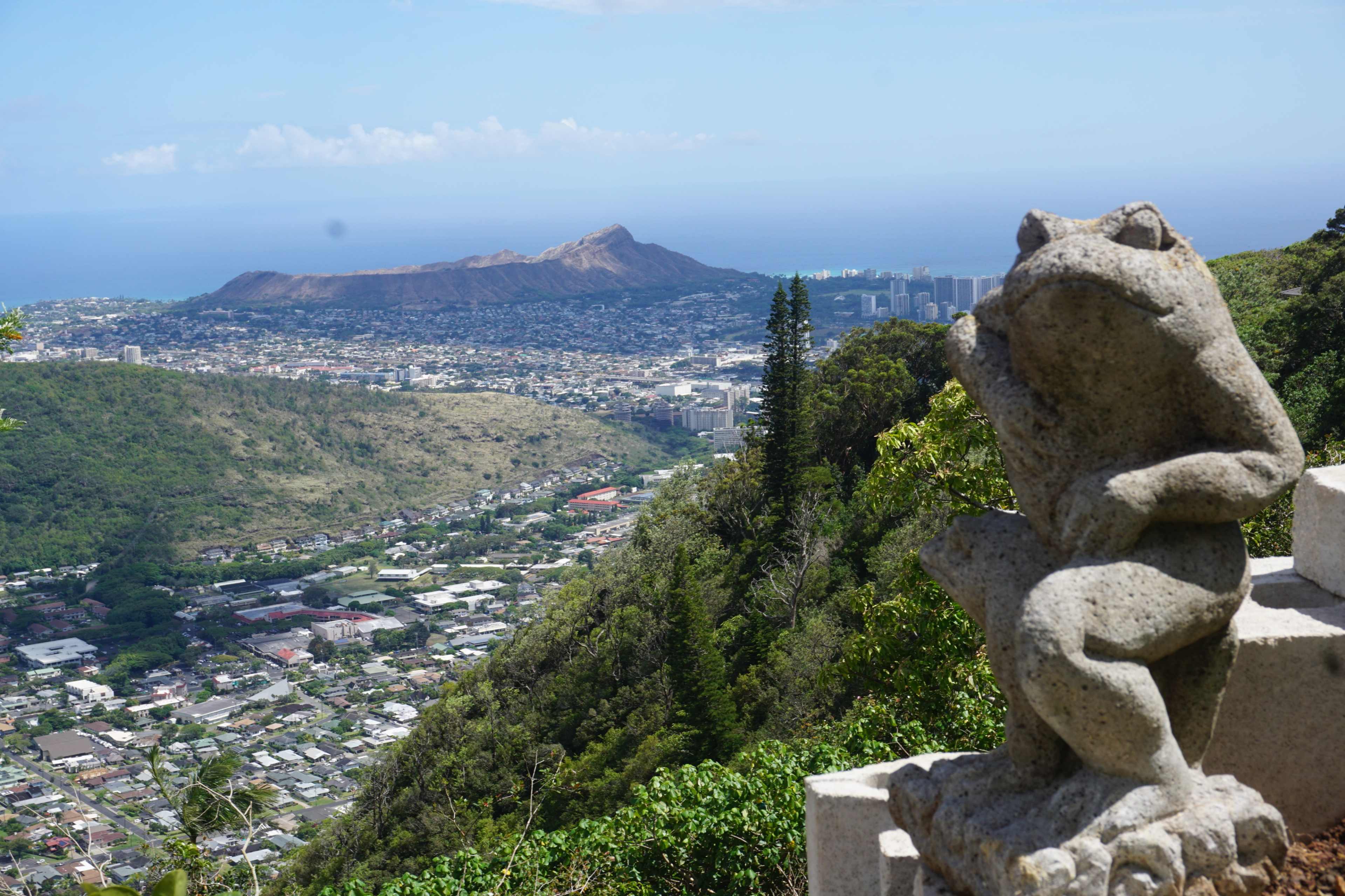 A stone frog statue overlooks a sprawling coastal city and Diamond Head in the distance.