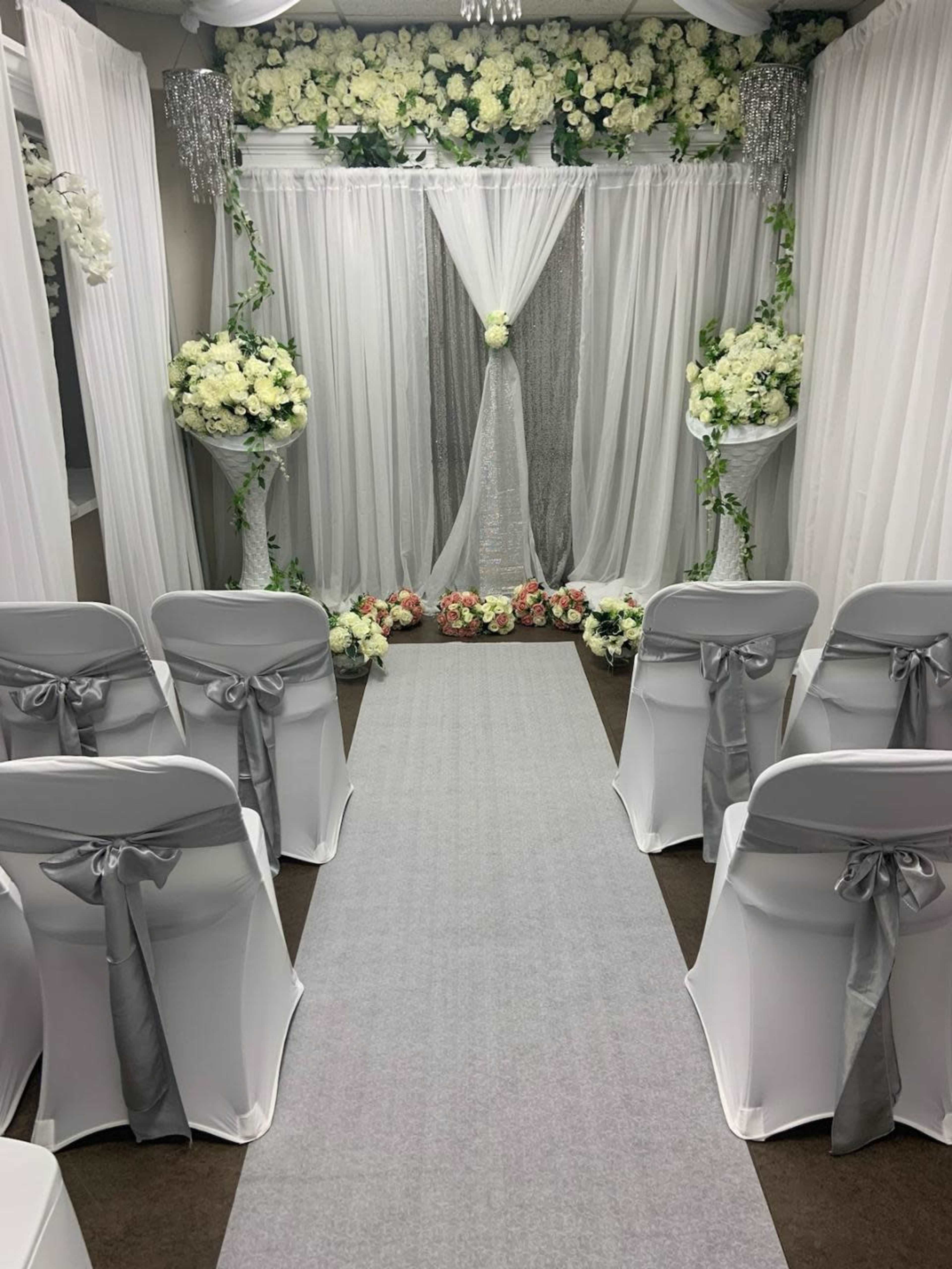The image shows a wedding ceremony setup with white chairs adorned with gray ribbons, a carpeted aisle, and a floral backdrop framed by draped curtains.
