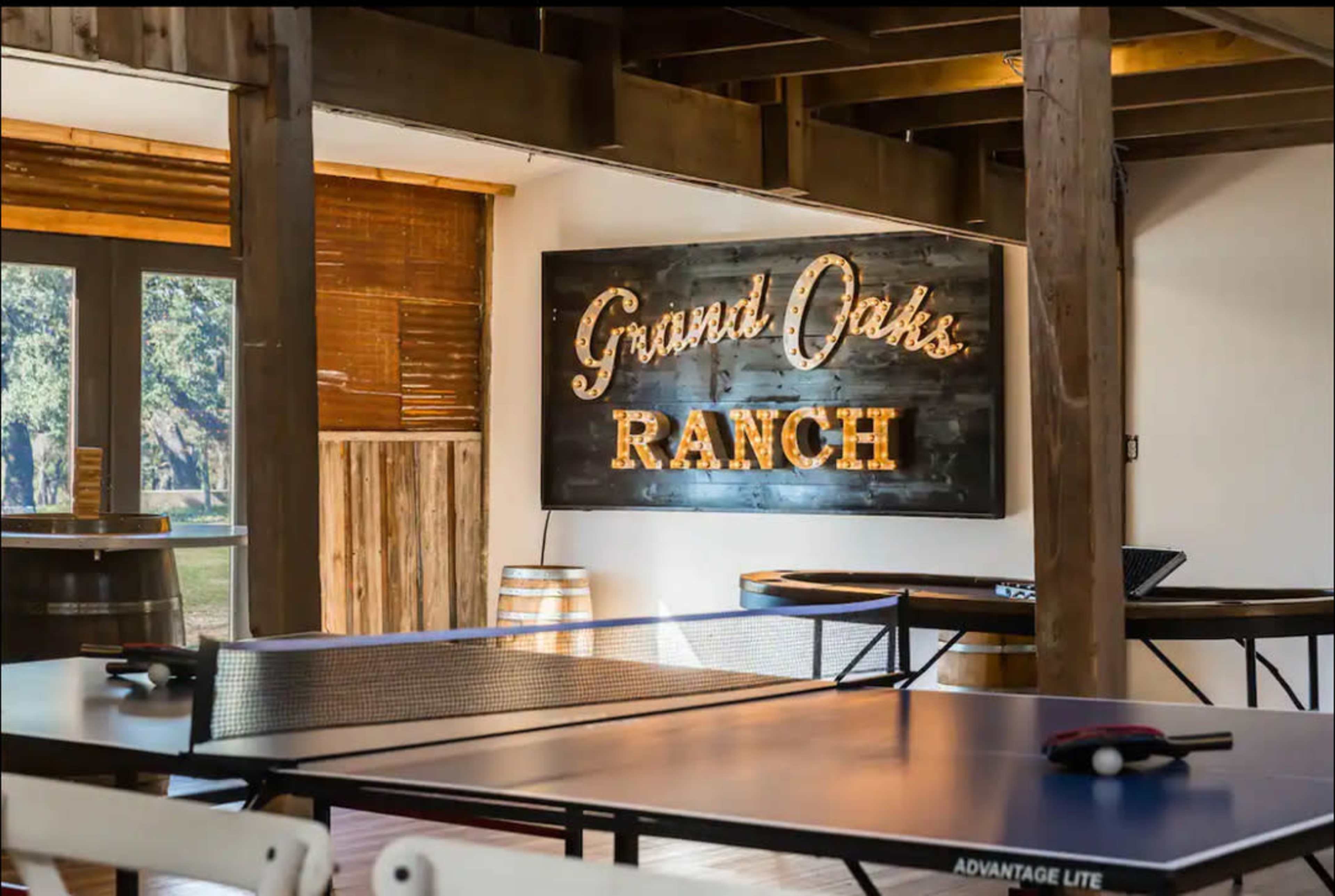 A lively indoor space featuring a ping pong table, wooden beams, and a bright sign that reads "Grand Oaks Ranch."