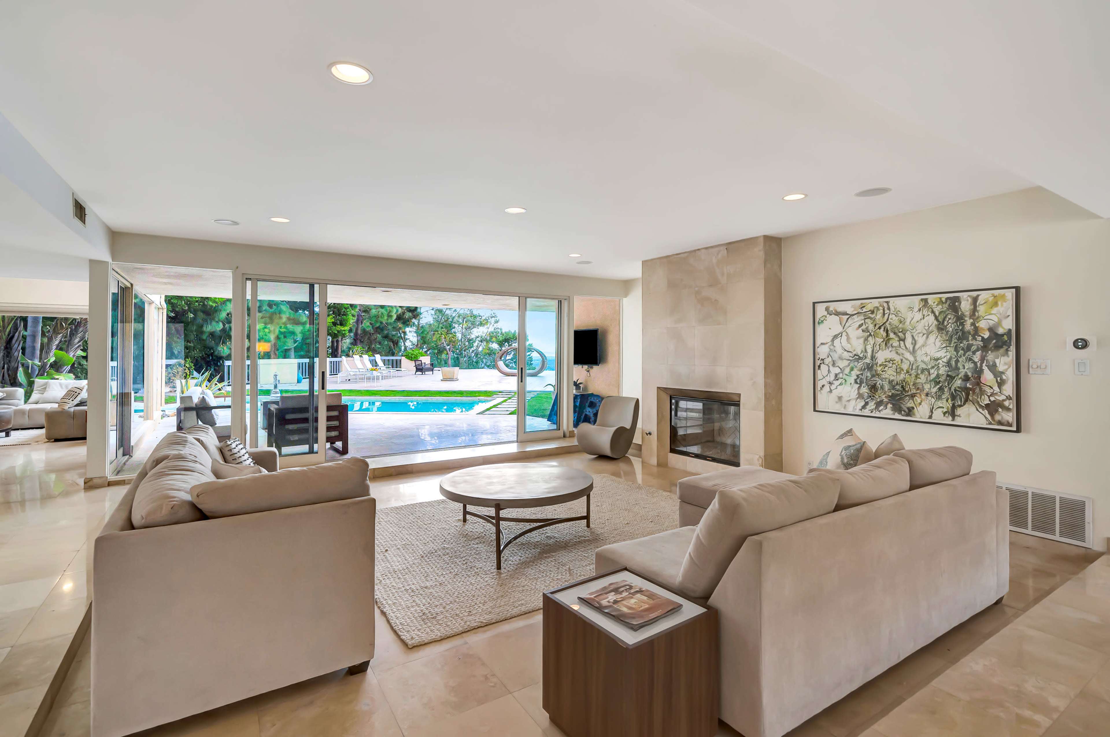 A modern living room features large windows overlooking a pool area, with beige sofas arranged around a round coffee table on a tiled floor.