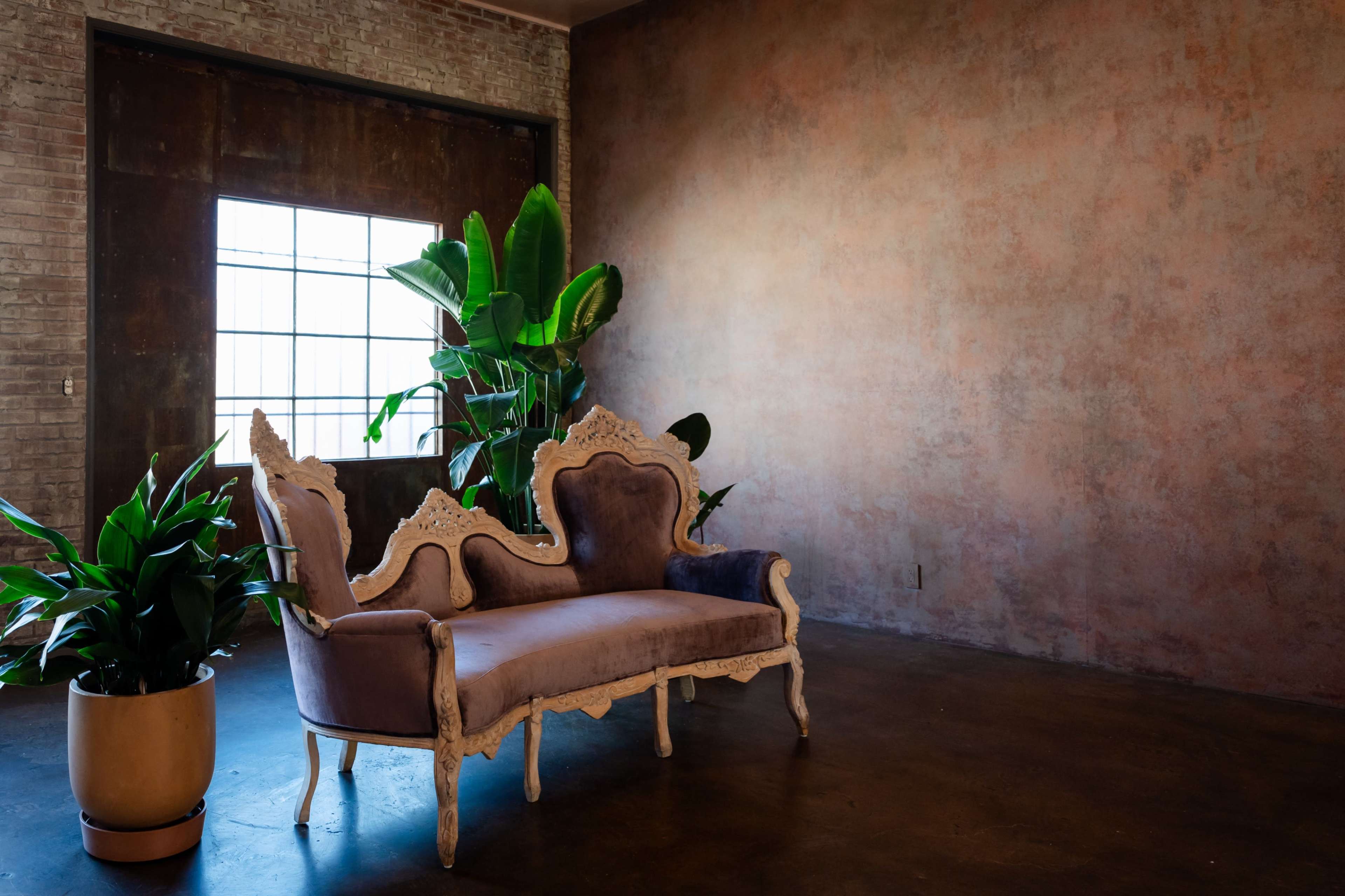 The image features a vintage-style sofa with ornate detailing positioned near a large window and accompanied by a potted plant in a spacious, brick-walled room.