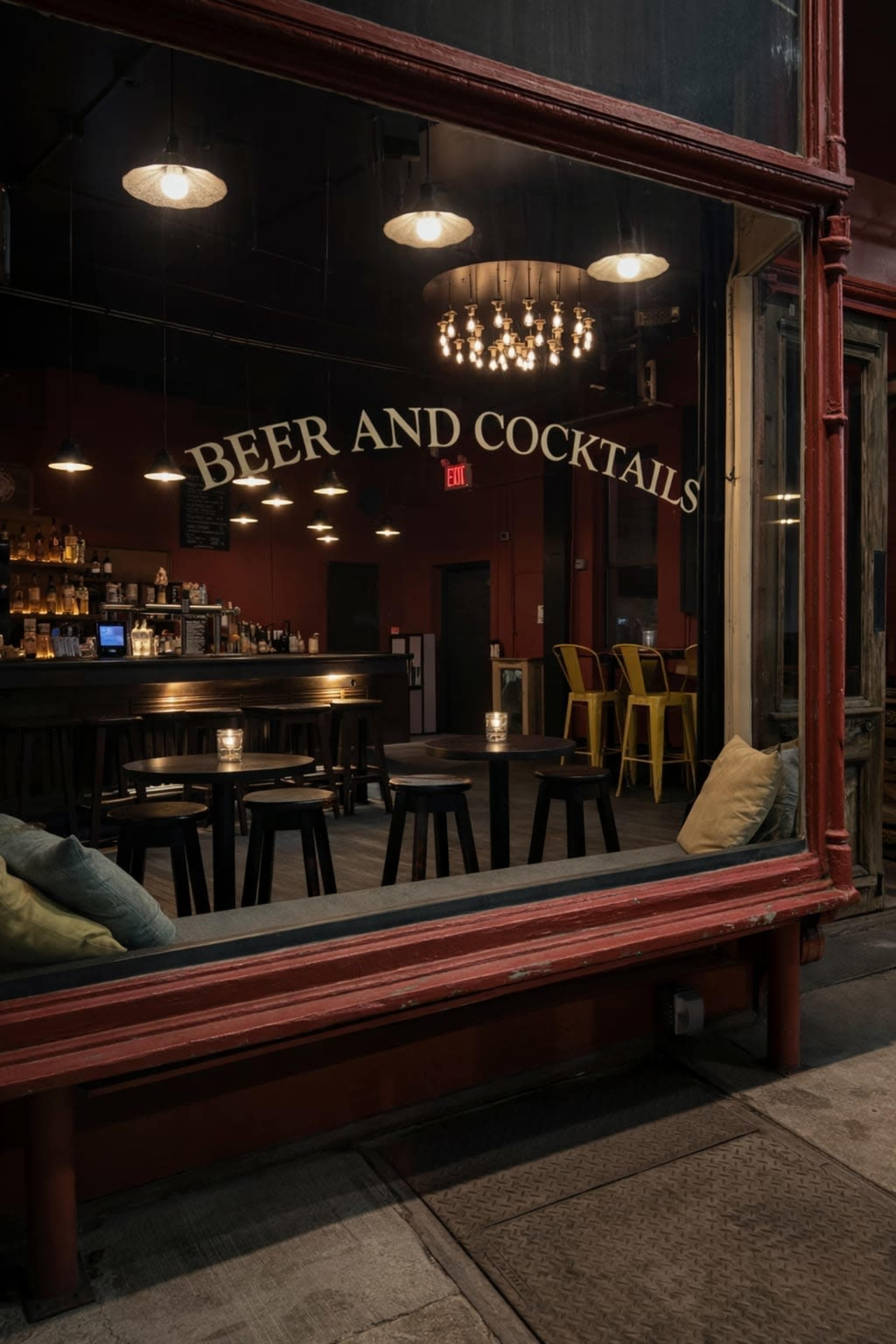 The dimly lit bar features a long counter with stools, illuminated by overhead lights and a central chandelier, with a sign stating "Beer and Cocktails" visible through the window.