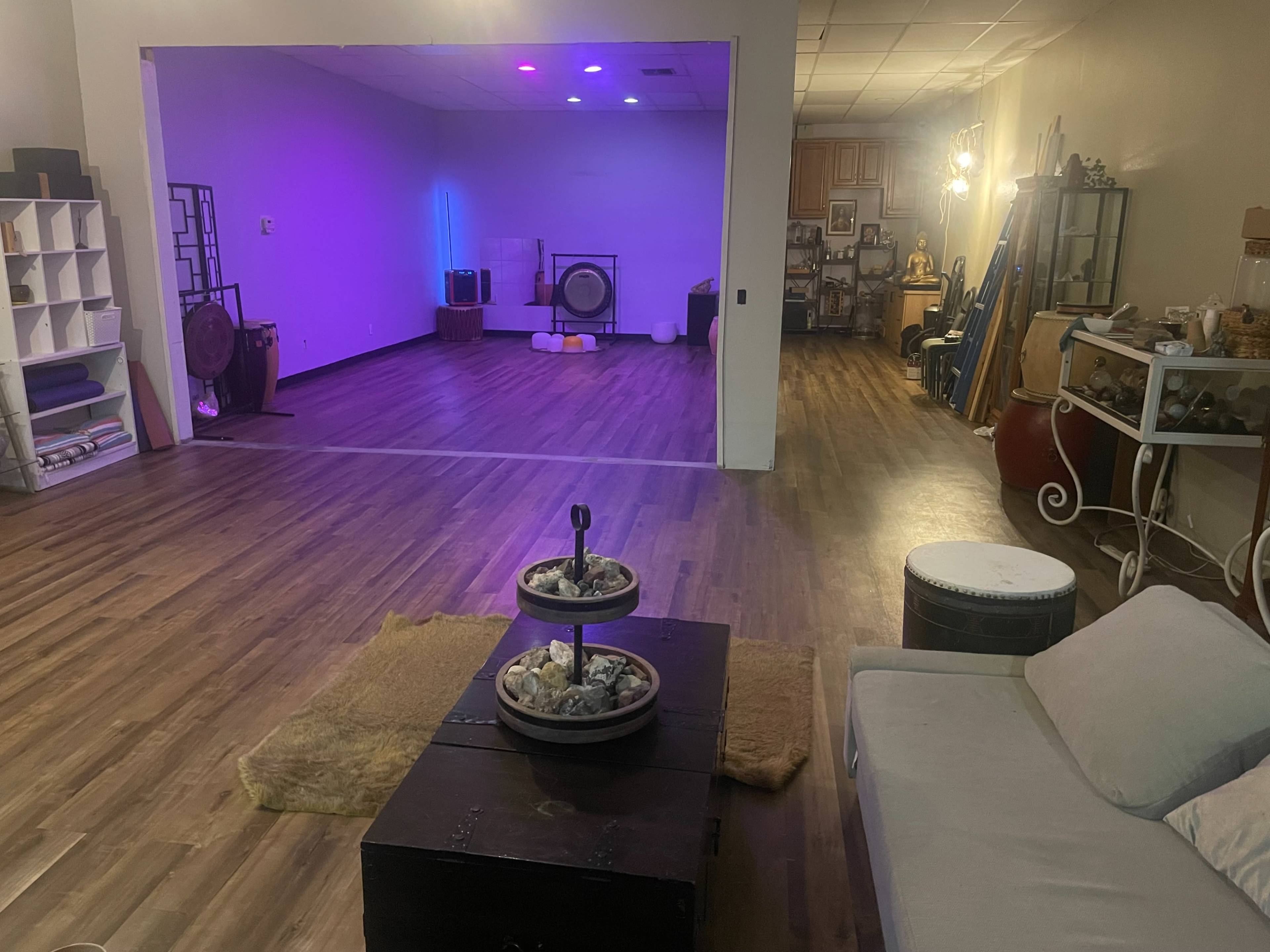 The image shows a spacious room with wooden flooring, a seating area featuring a low table and a couch, and a brightly lit area in the background with various decorative items and instruments.