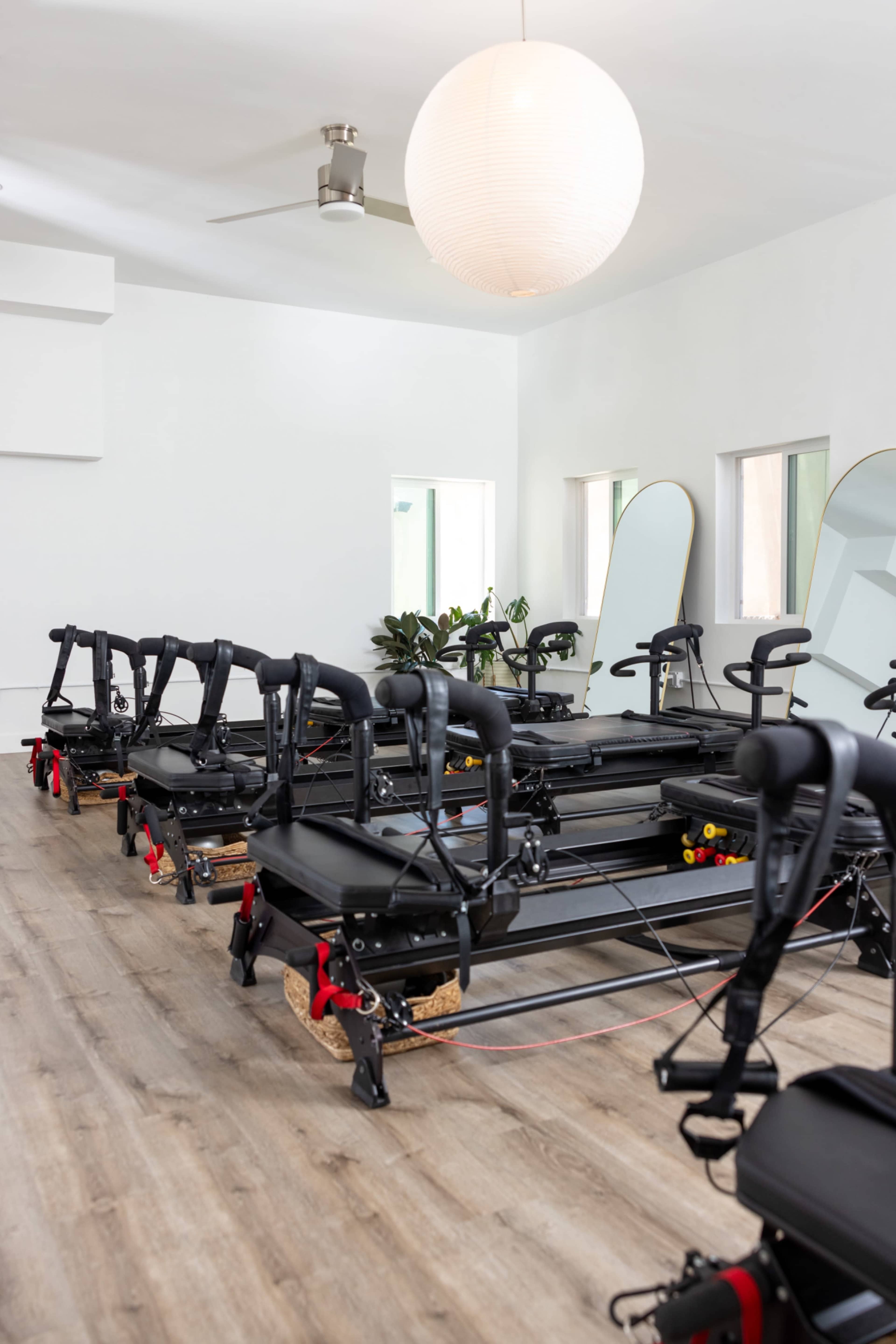 The image shows a bright, modern exercise studio equipped with several reformer machines lined up neatly on a wooden floor.
