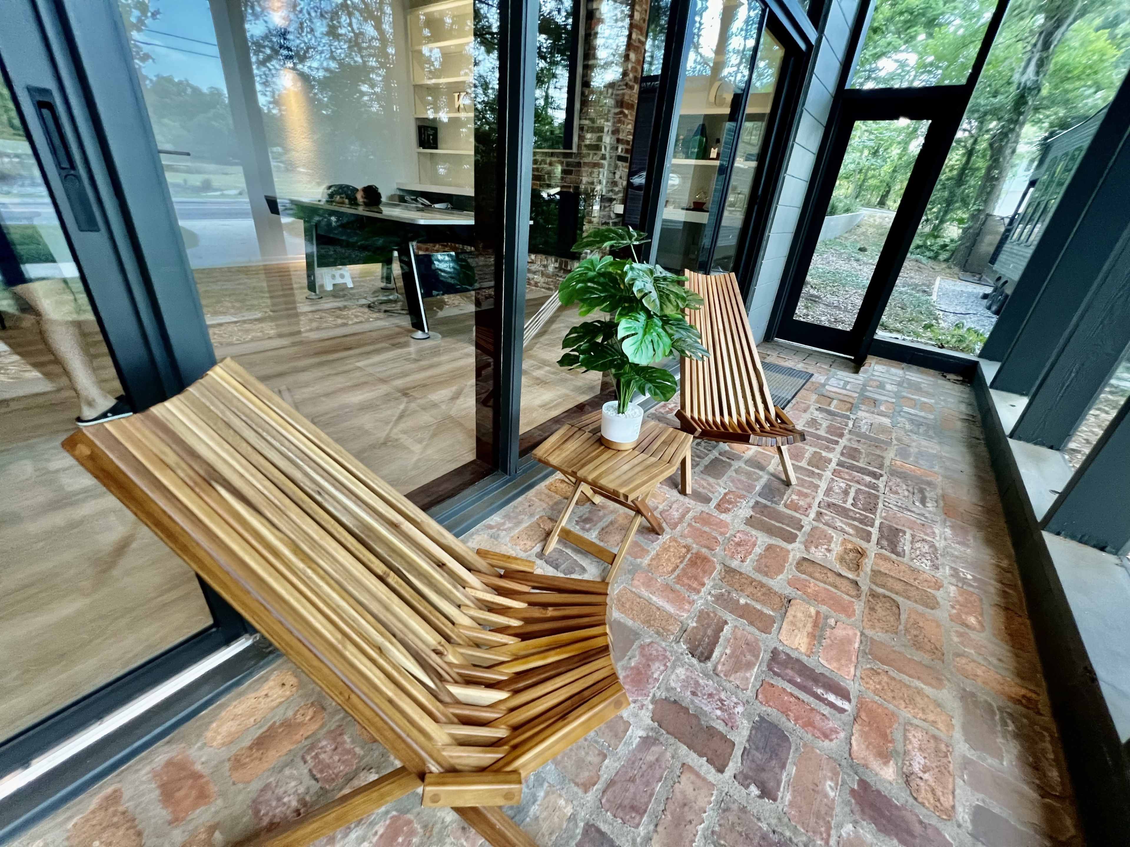 The image shows a porch area featuring two wooden chairs and a small table with a potted plant, complemented by a brick floor and large glass windows.