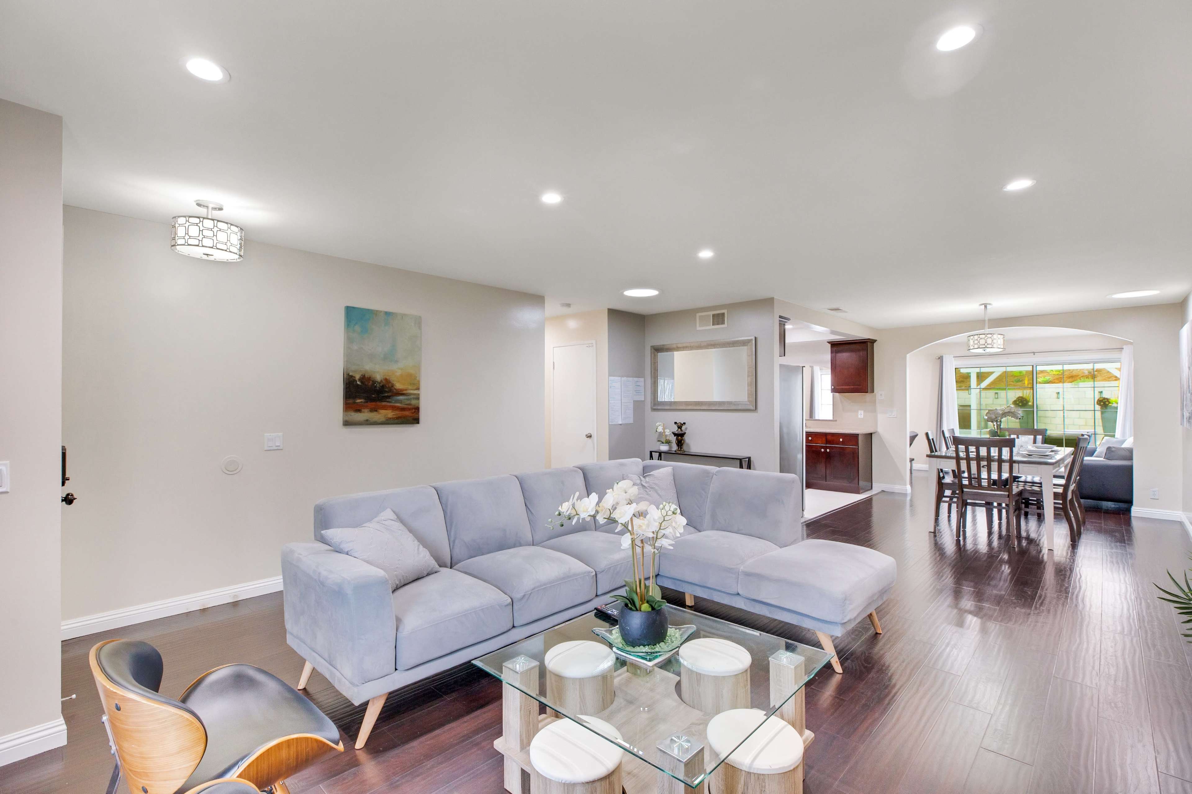 A modern living room features a light gray sectional sofa, a glass coffee table with decorative items, and an open layout leading to a dining area and kitchen.