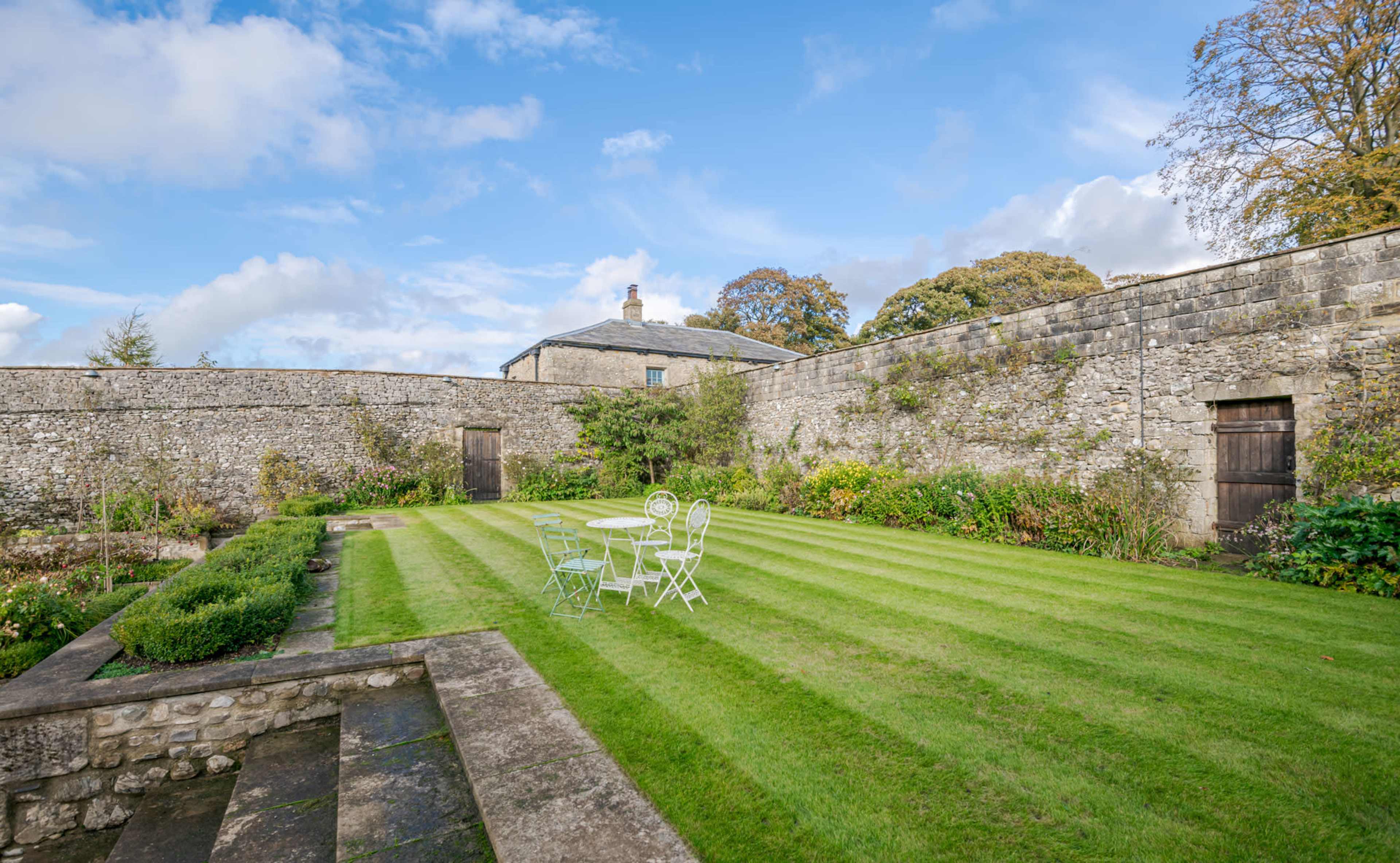 A well-maintained garden features neatly striped grass and a small table set with chairs, enclosed by a stone wall.