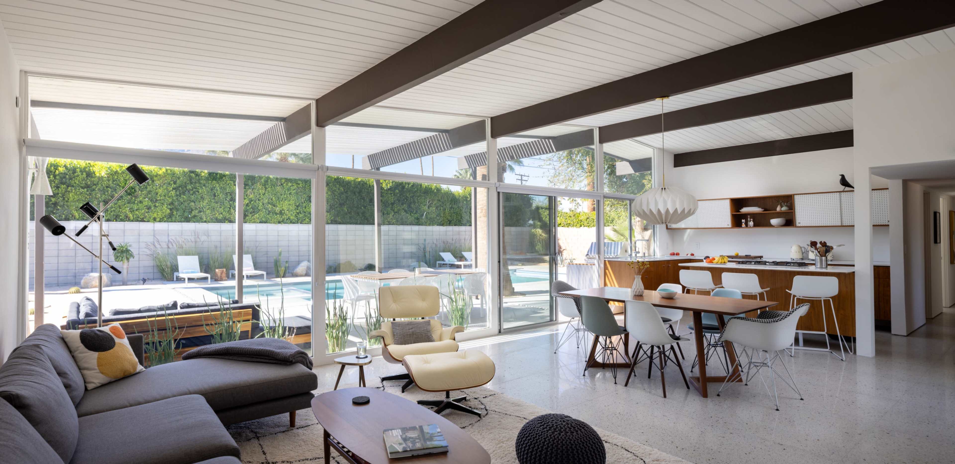 The image shows a modern open-concept living room and kitchen area with large windows overlooking a backyard pool.