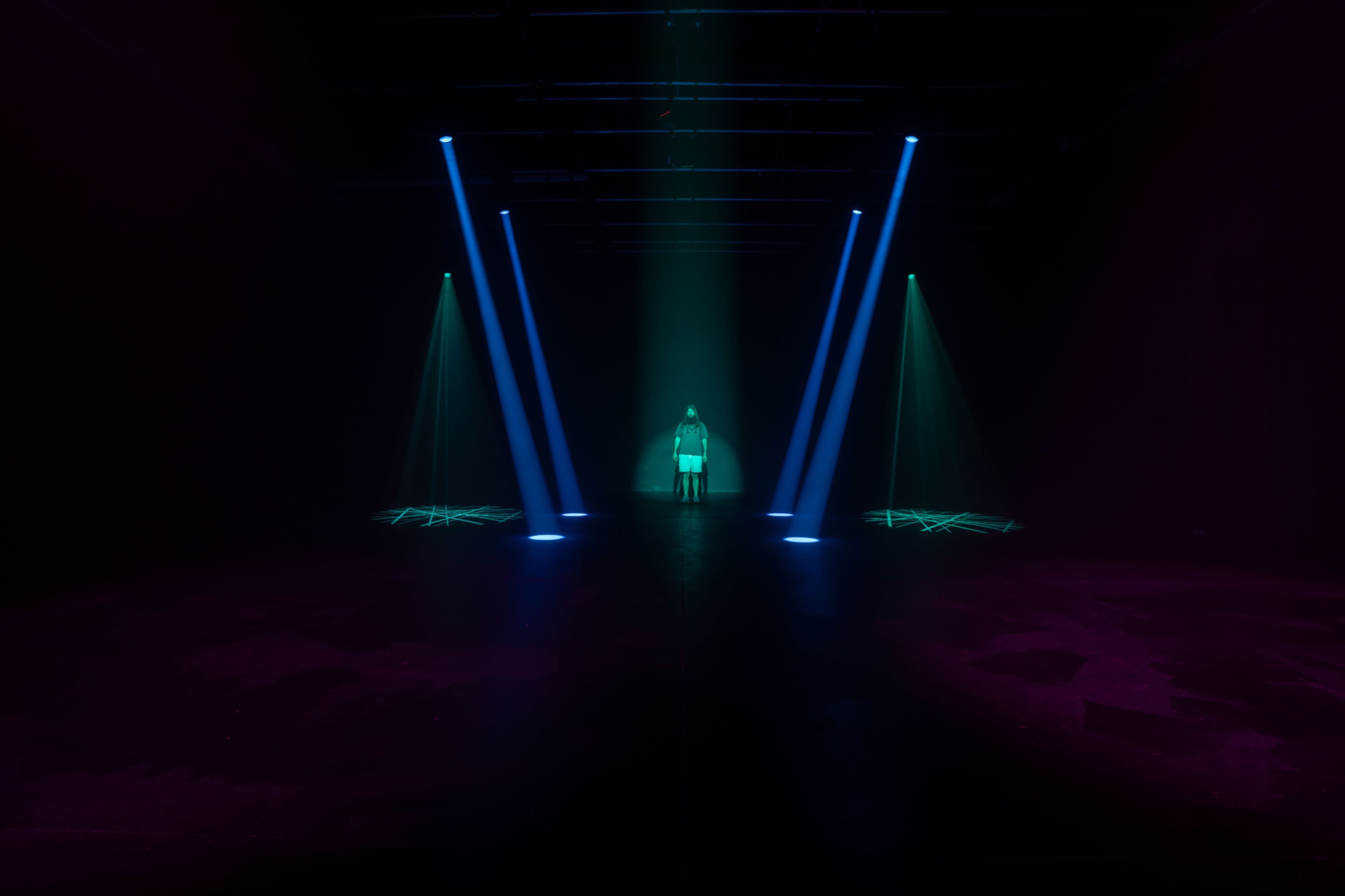 A figure stands in a dark space illuminated by vibrant beams of light in blue and green, casting shapes on the floor.