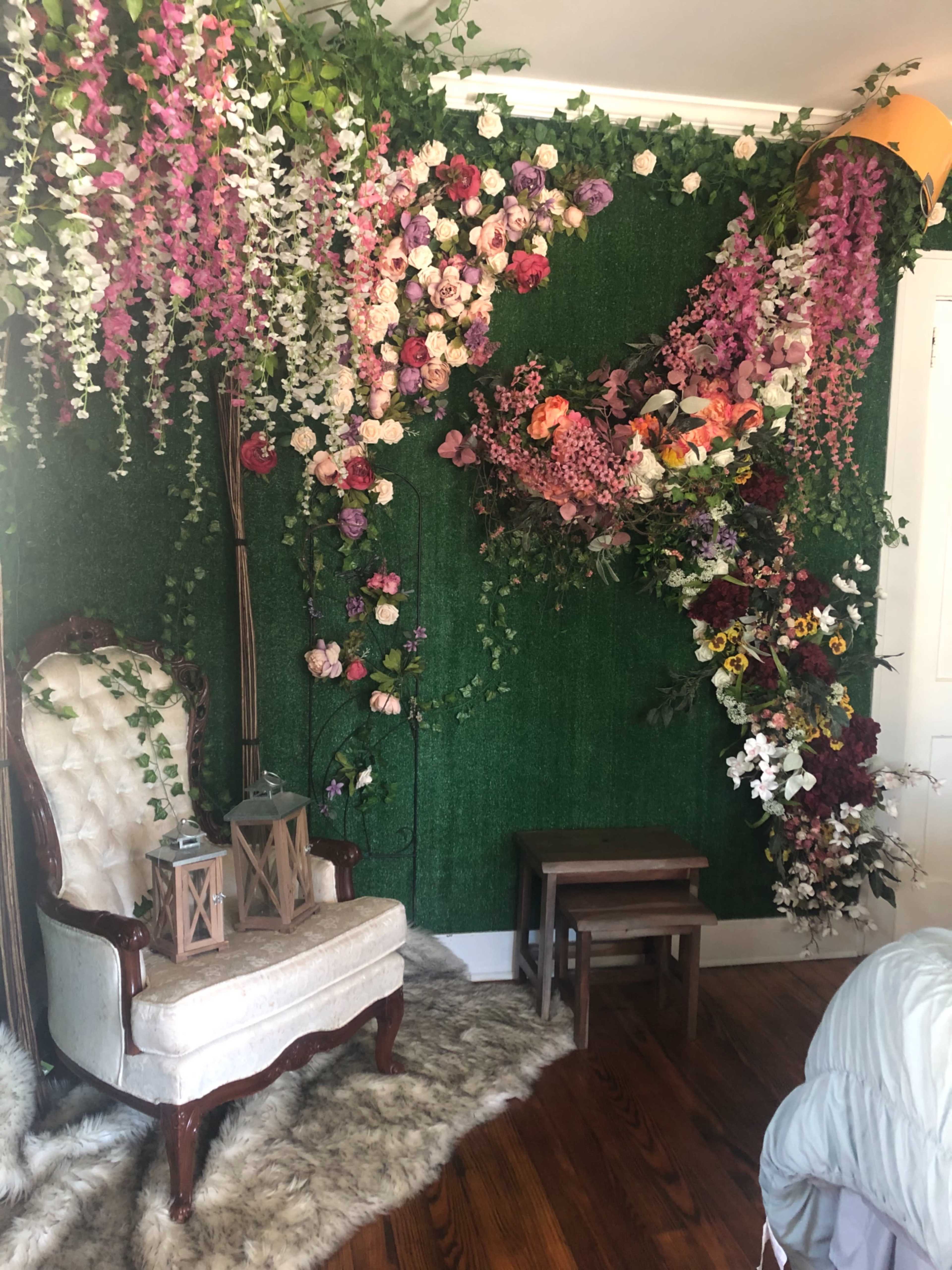 A decorated corner of a room featuring an ornate white chair, a small wooden table, and a vibrant floral wall display with greenery.