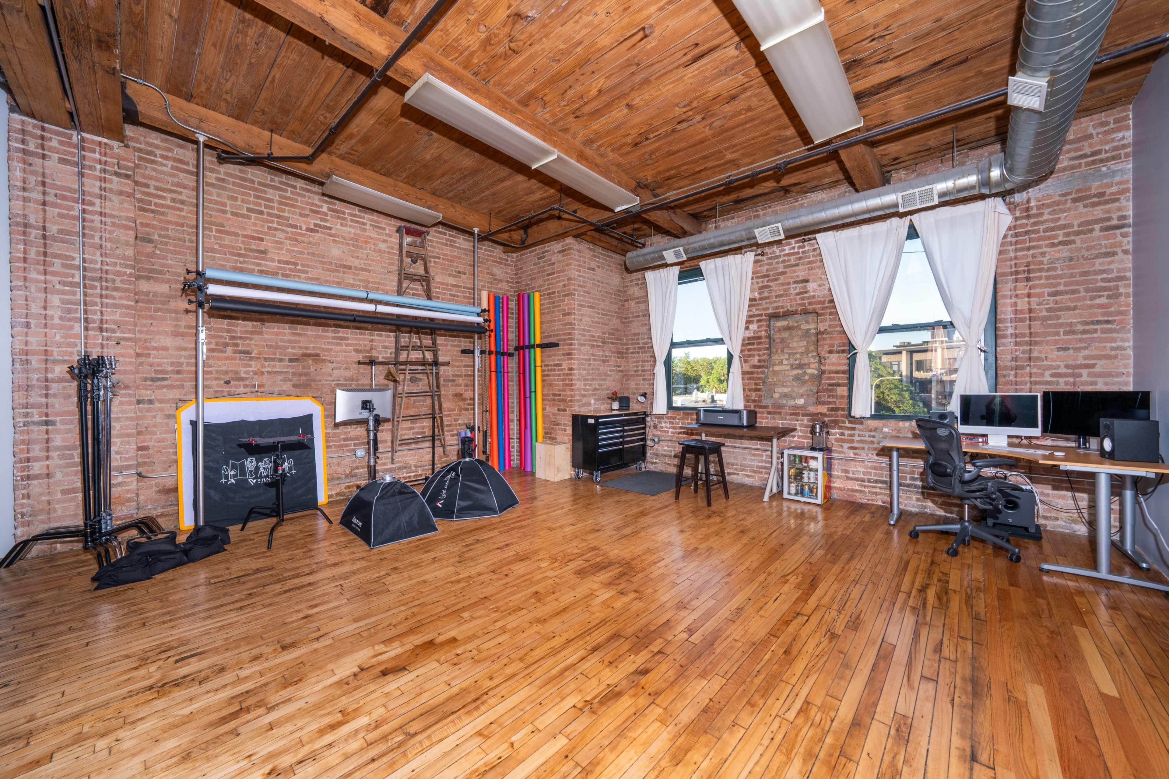 The image shows a spacious photography studio with wooden flooring, exposed brick walls, and various equipment including backdrops, lighting stands, and a desk with computer monitors.
